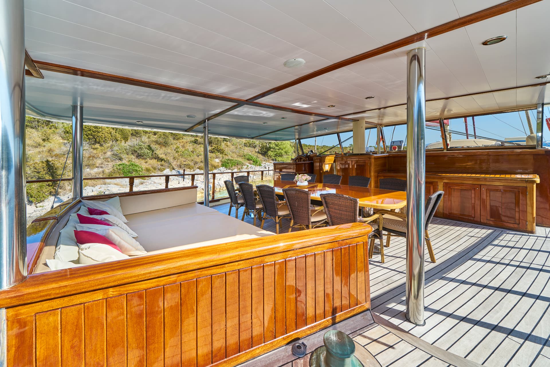Yacht deck with dining area and sunbed near a rocky, scrub-covered coastline.