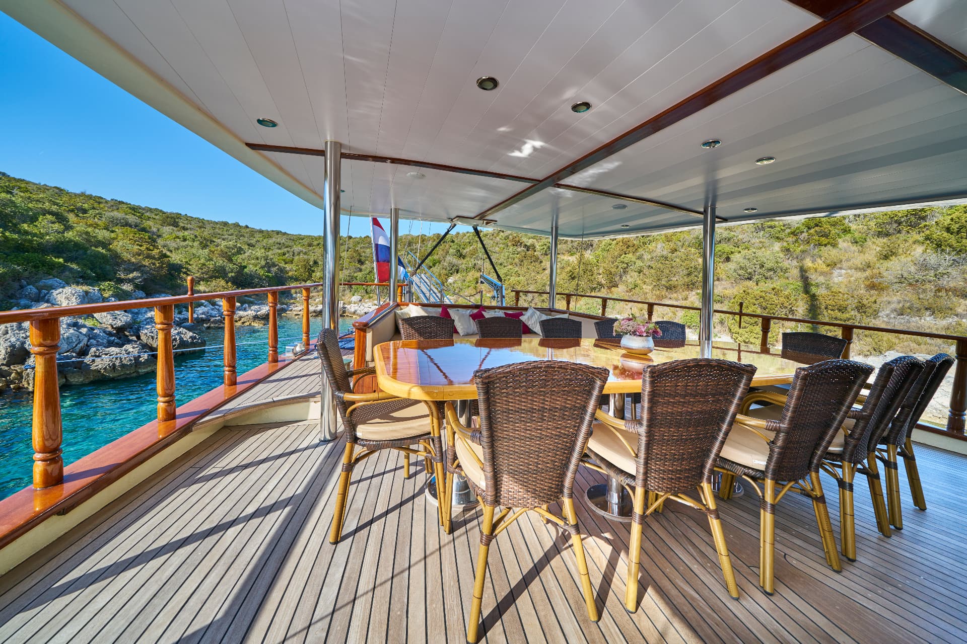Dining area on a yacht deck with wicker chairs next to turquoise coastal water and green hills.