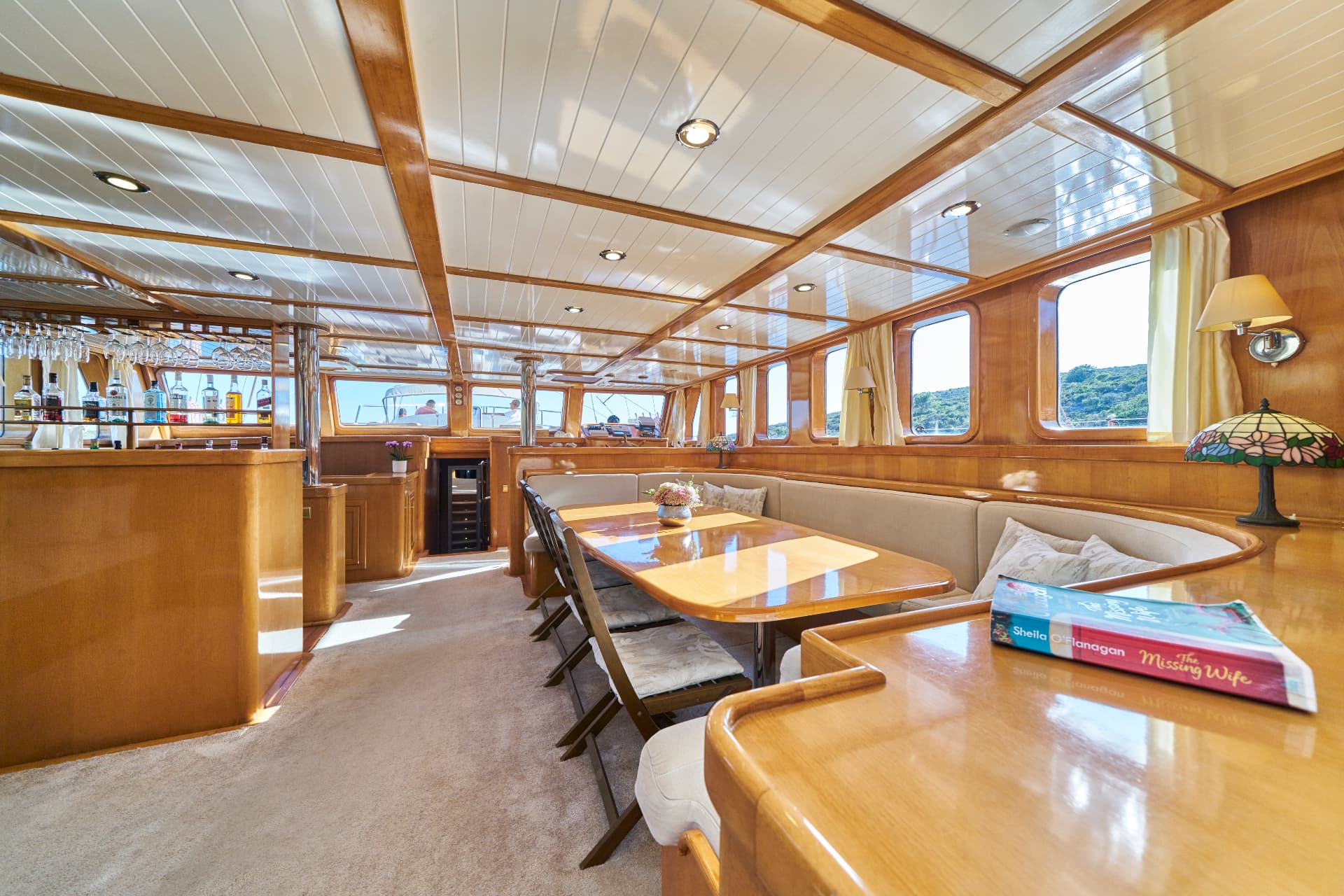 Yacht interior with wood paneling, dining table, bar area, and view of green coastline.