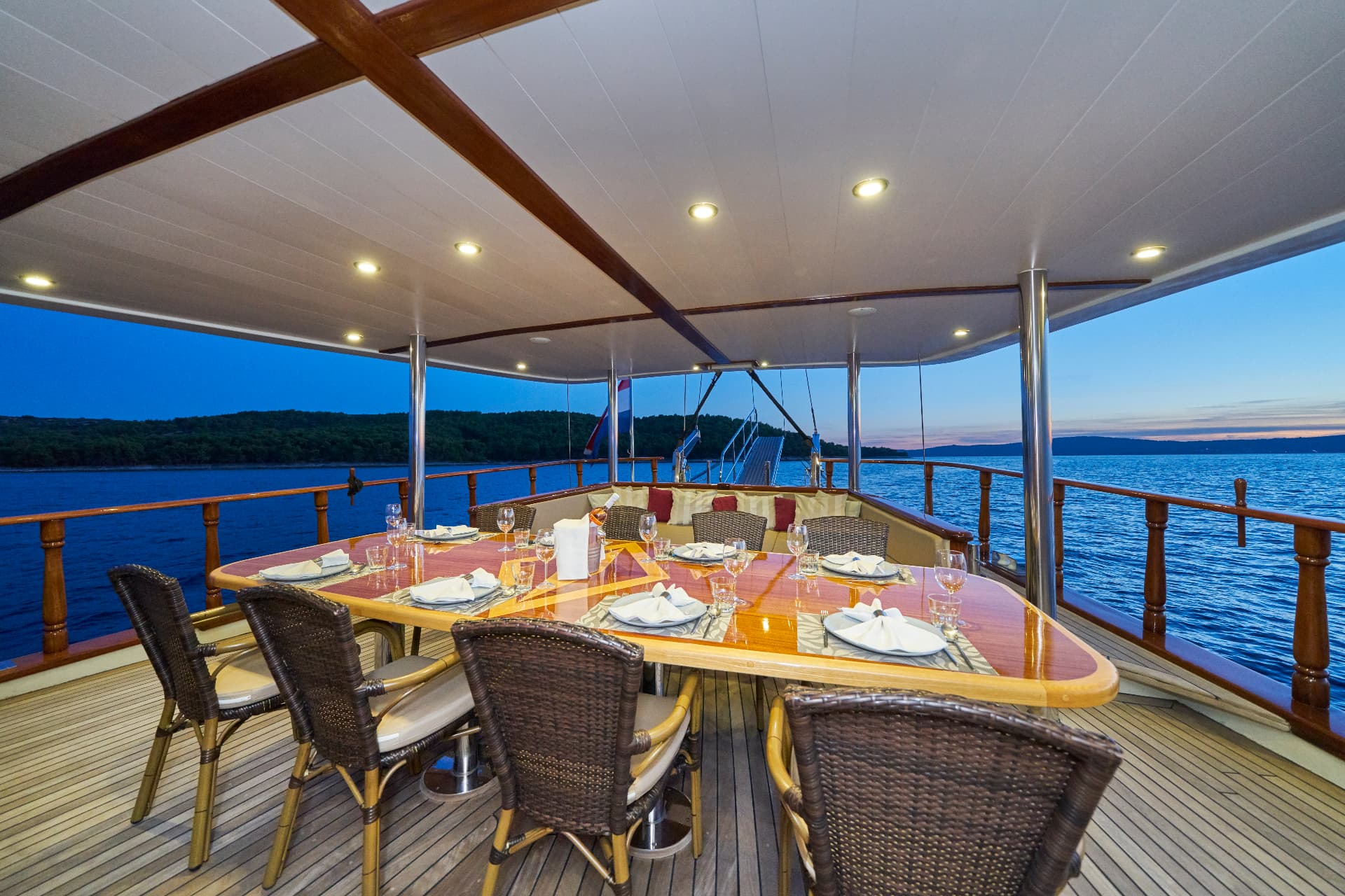 Outdoor dining area on a boat deck with set table overlooking dark water and forested coastline at dusk.