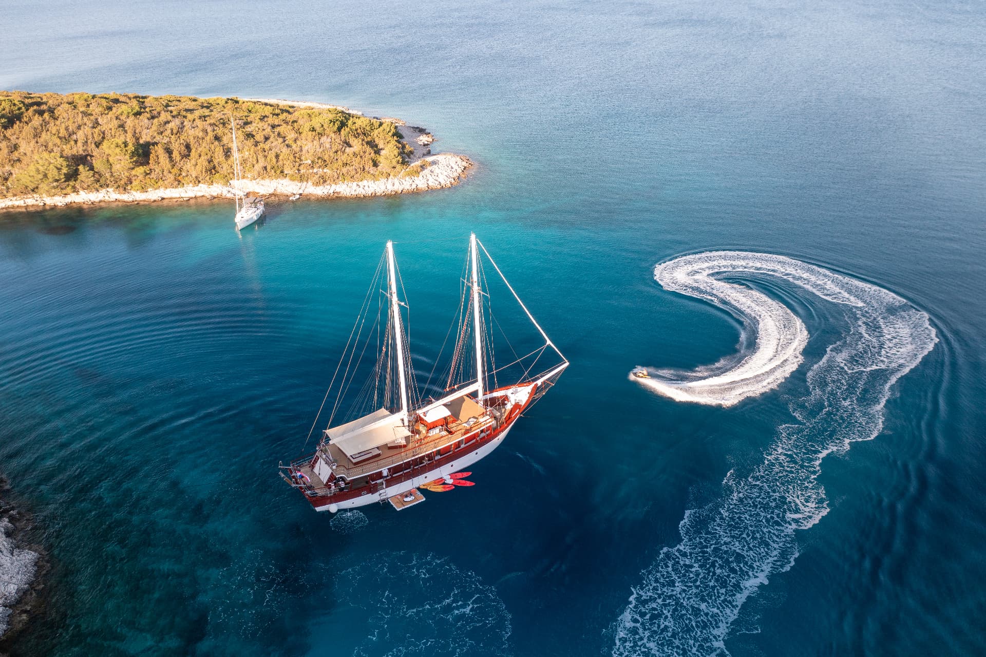 Large sailboat anchored near a wooded island while a jet ski leaves a white wake on turquoise sea.