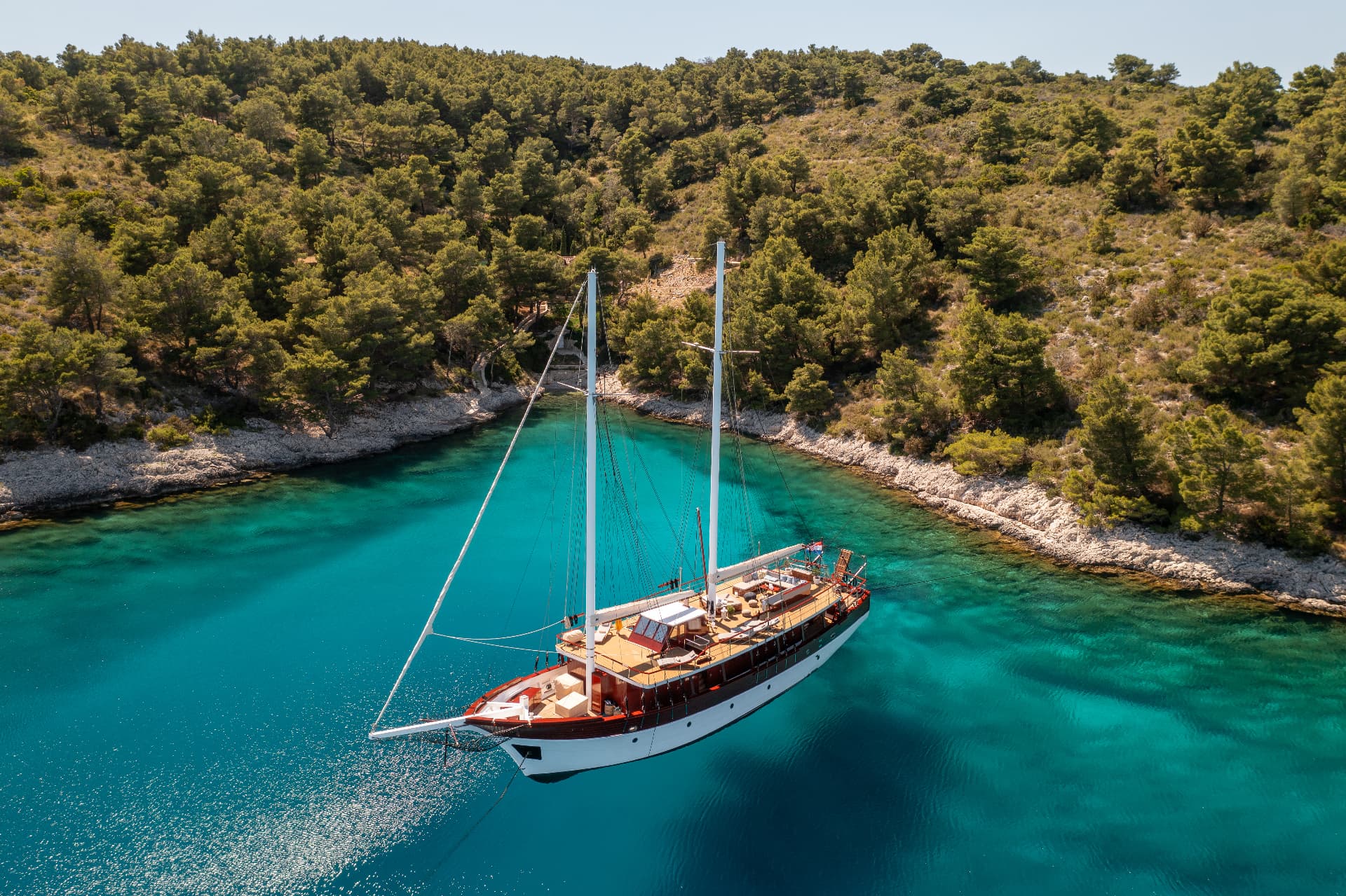 Large wooden sailboat anchored in turquoise cove surrounded by pine-covered rocky coastline.