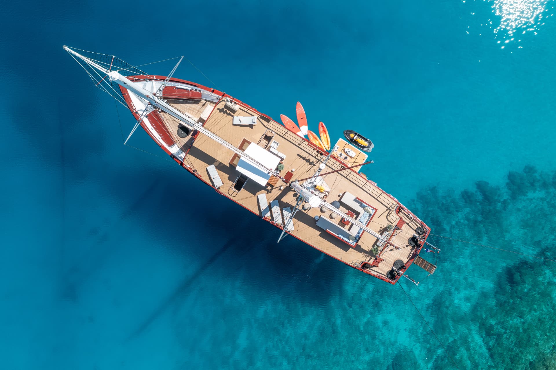 Aerial view of large wooden yacht anchored in clear turquoise coastal waters with paddleboards on deck.