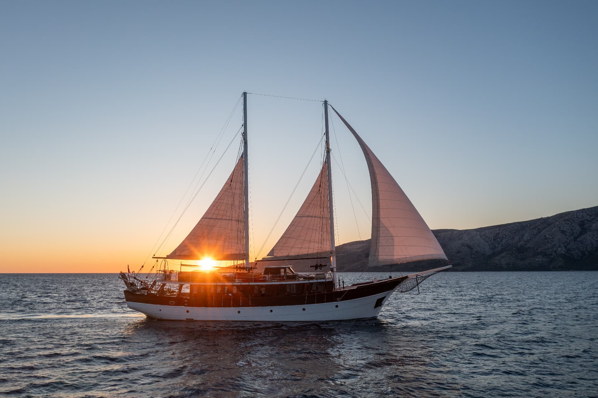 Two-masted sailboat cruising on the sea at sunset near a dark, hilly coastline.