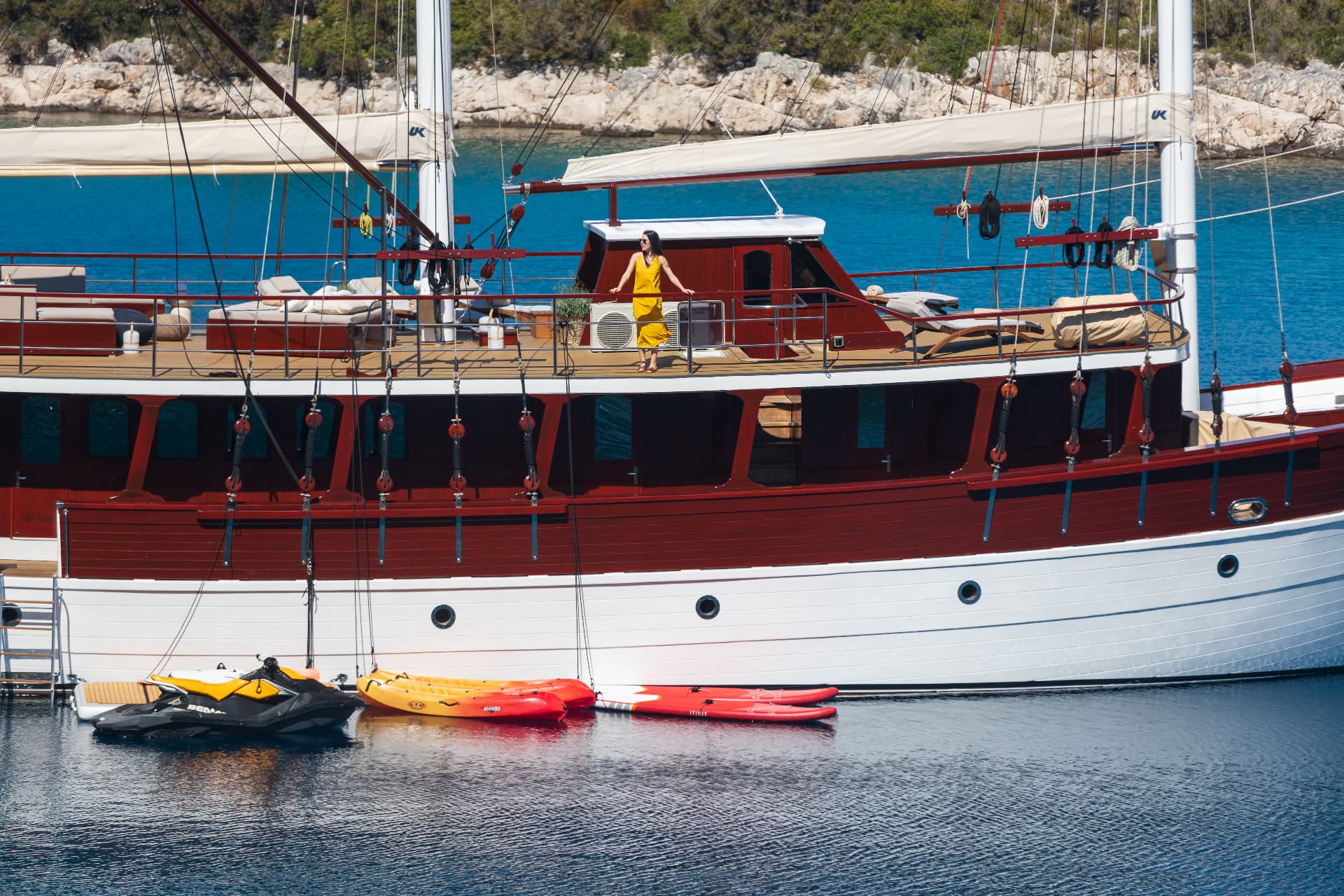 Large wooden yacht docked near rocky, green coastline with jet ski and kayaks floating nearby.
