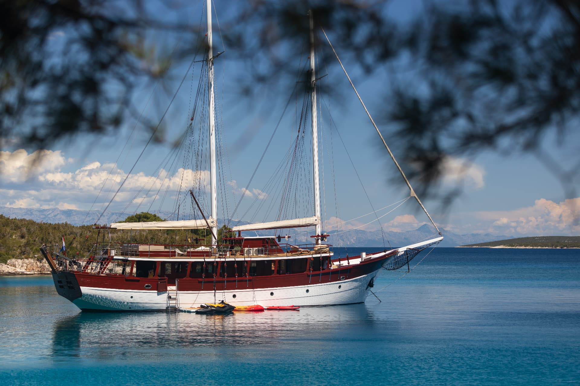 Large wooden sailboat anchored in turquoise coastal waters with mountains in background