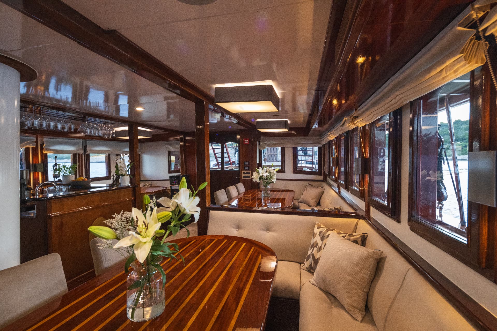 Interior dining area of a yacht with polished wood tables and white lilies, looking out to sea.