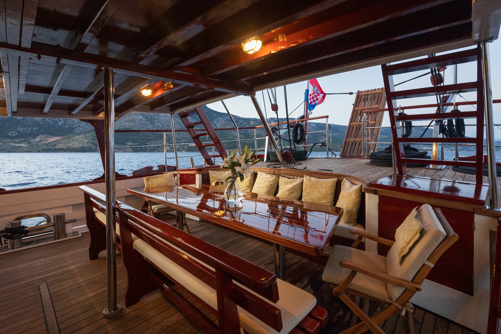 Boat deck dining area with glossy wood table, yellow cushions, and Croatian flag near mountains.