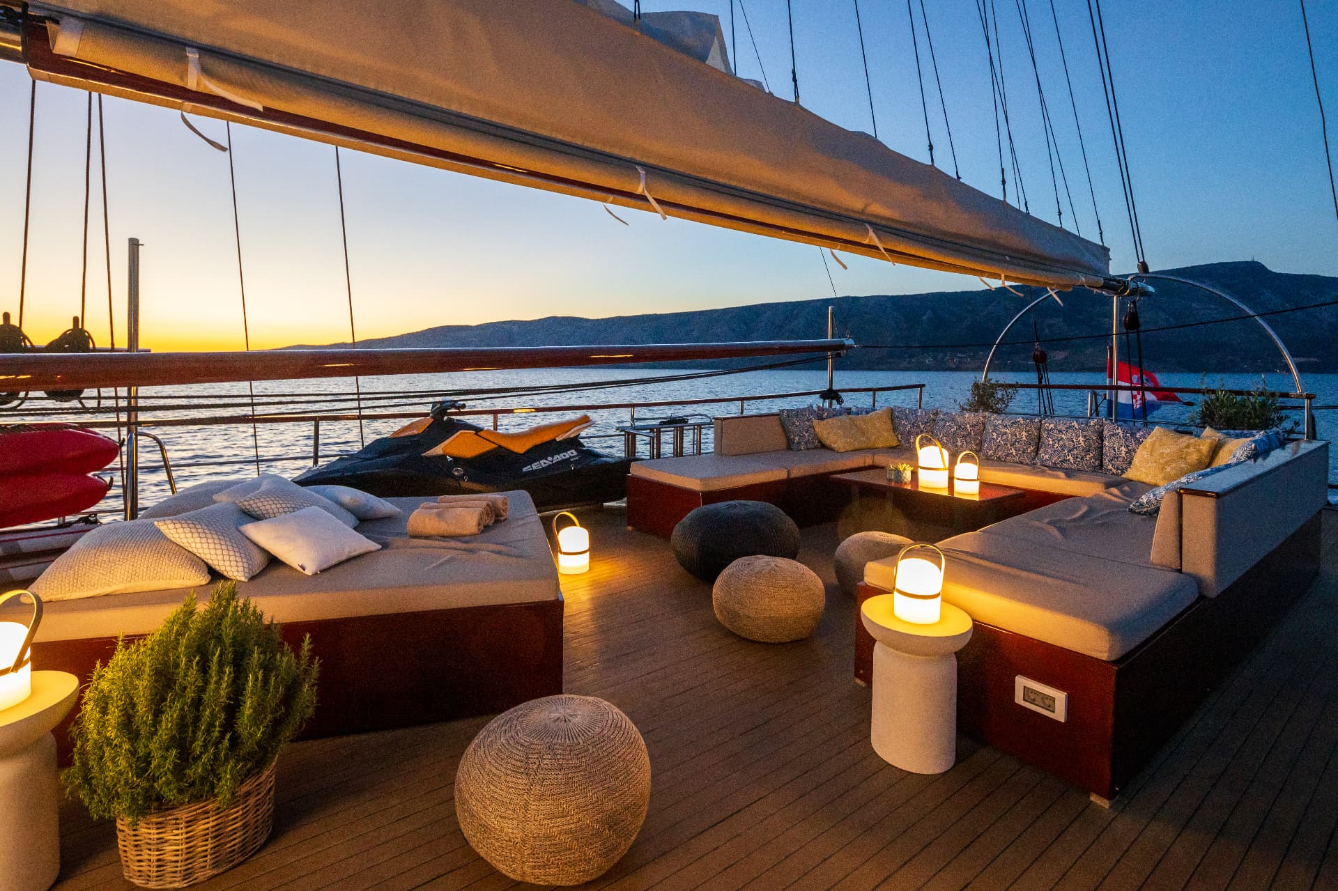 Yacht deck lounge area with cushions and lanterns at sunset over the sea and mountains.