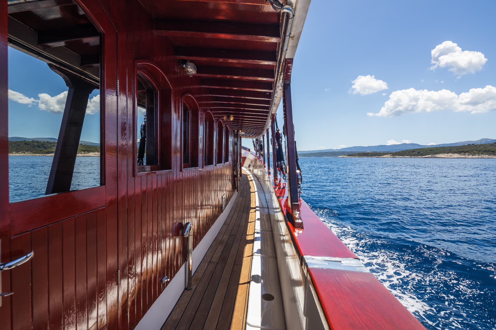 Wooden boat deck alongside glossy red cabin structure moving across blue sea near coastline.