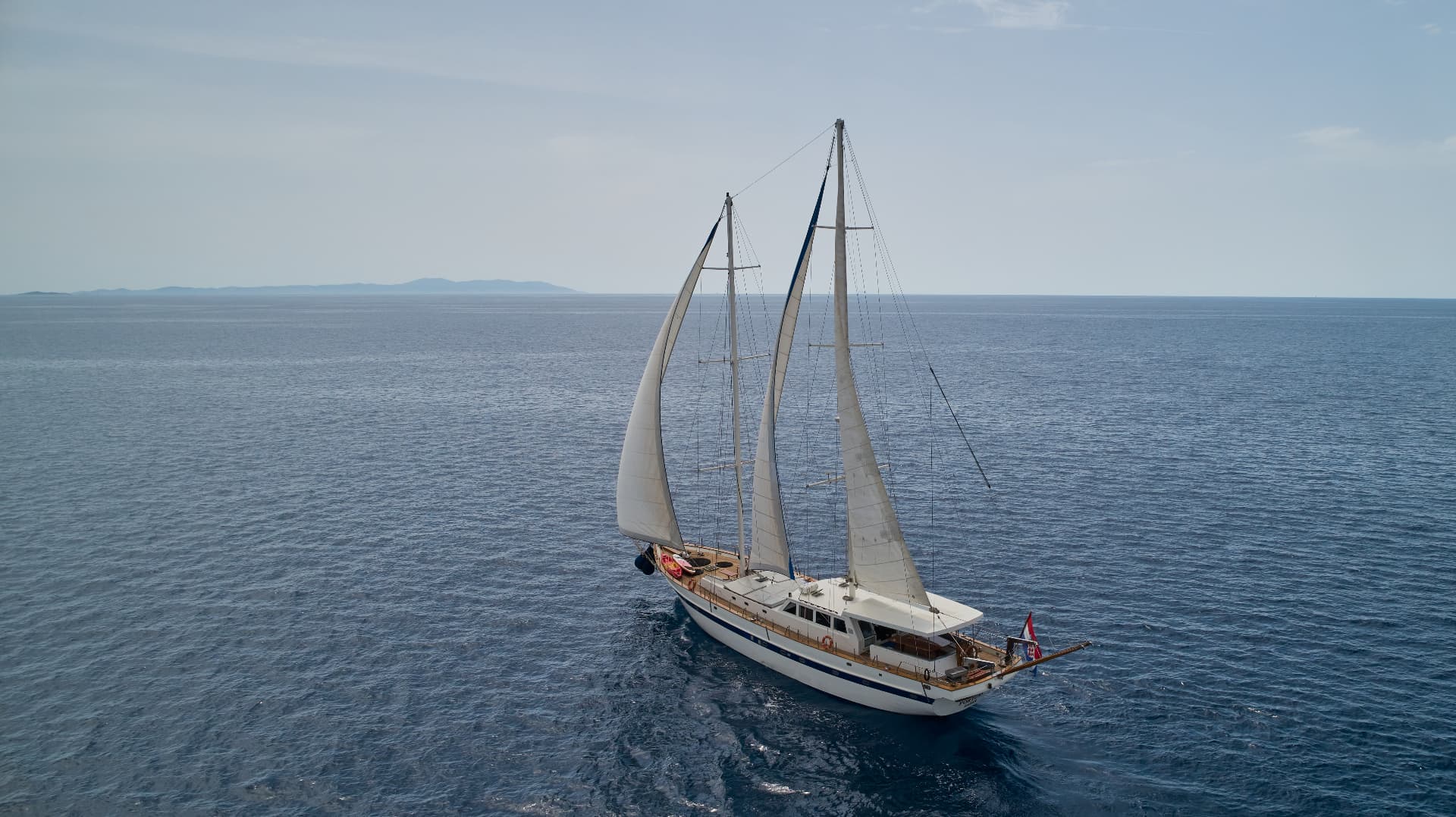 Large white sailboat with sails up cruising on deep blue sea with distant islands visible.
