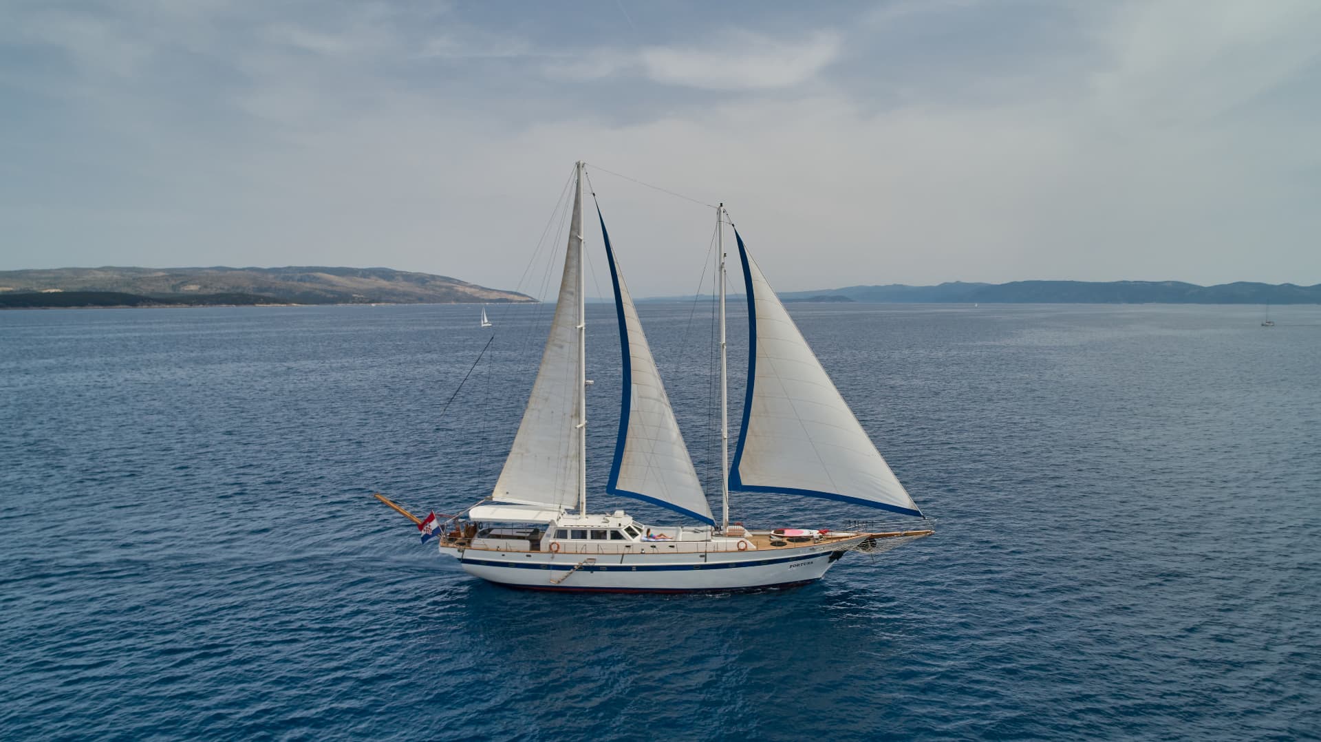 Large sailboat named Fortuna with white and blue sails on deep blue sea near hilly coastline.