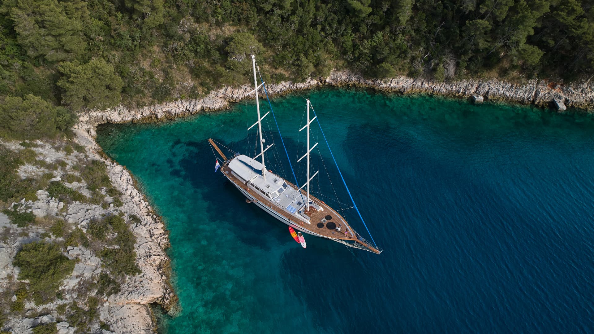 Large sailboat anchored in turquoise cove near rocky, forested coastline from above.