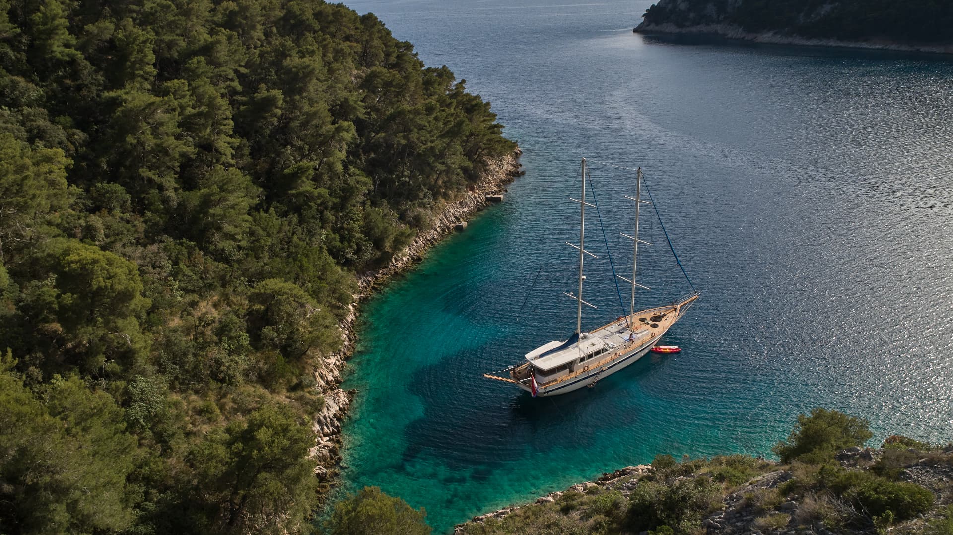 Large sailboat anchored in turquoise cove next to densely forested rocky coastline.