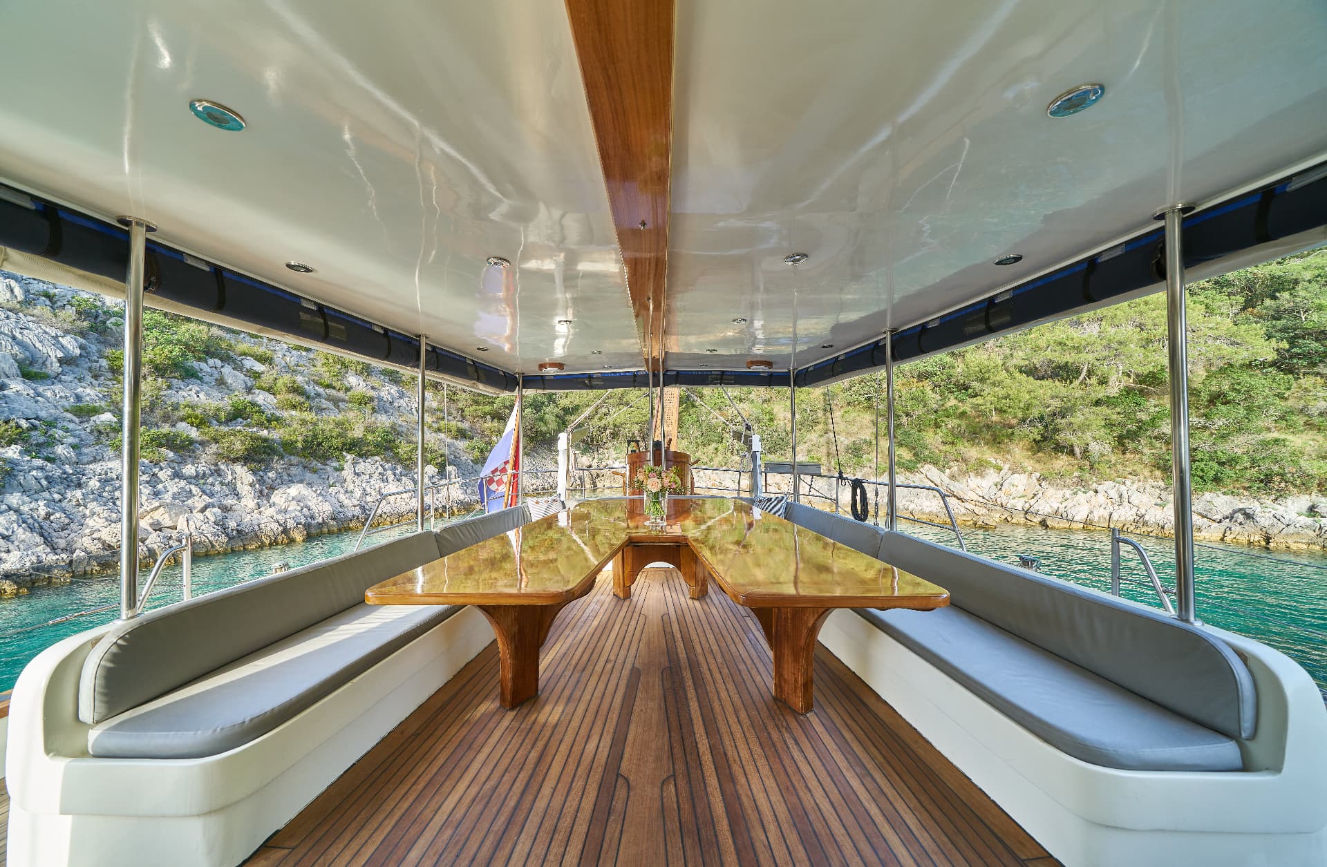 Boat deck with wooden table and seating next to rocky, green coastline.