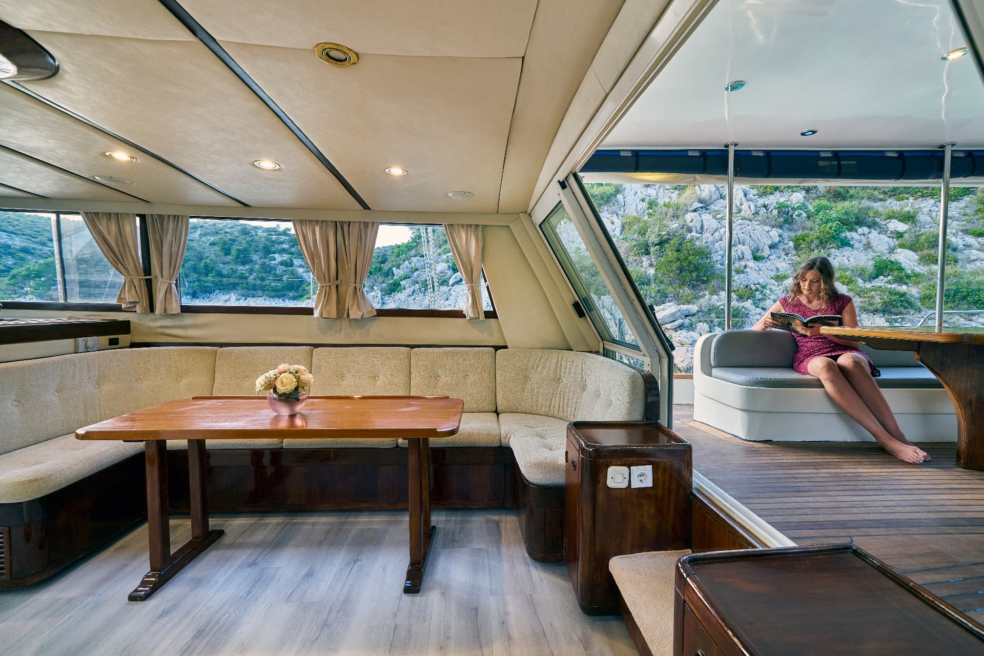 Woman reading on boat deck near rocky, green coastline visible through windows