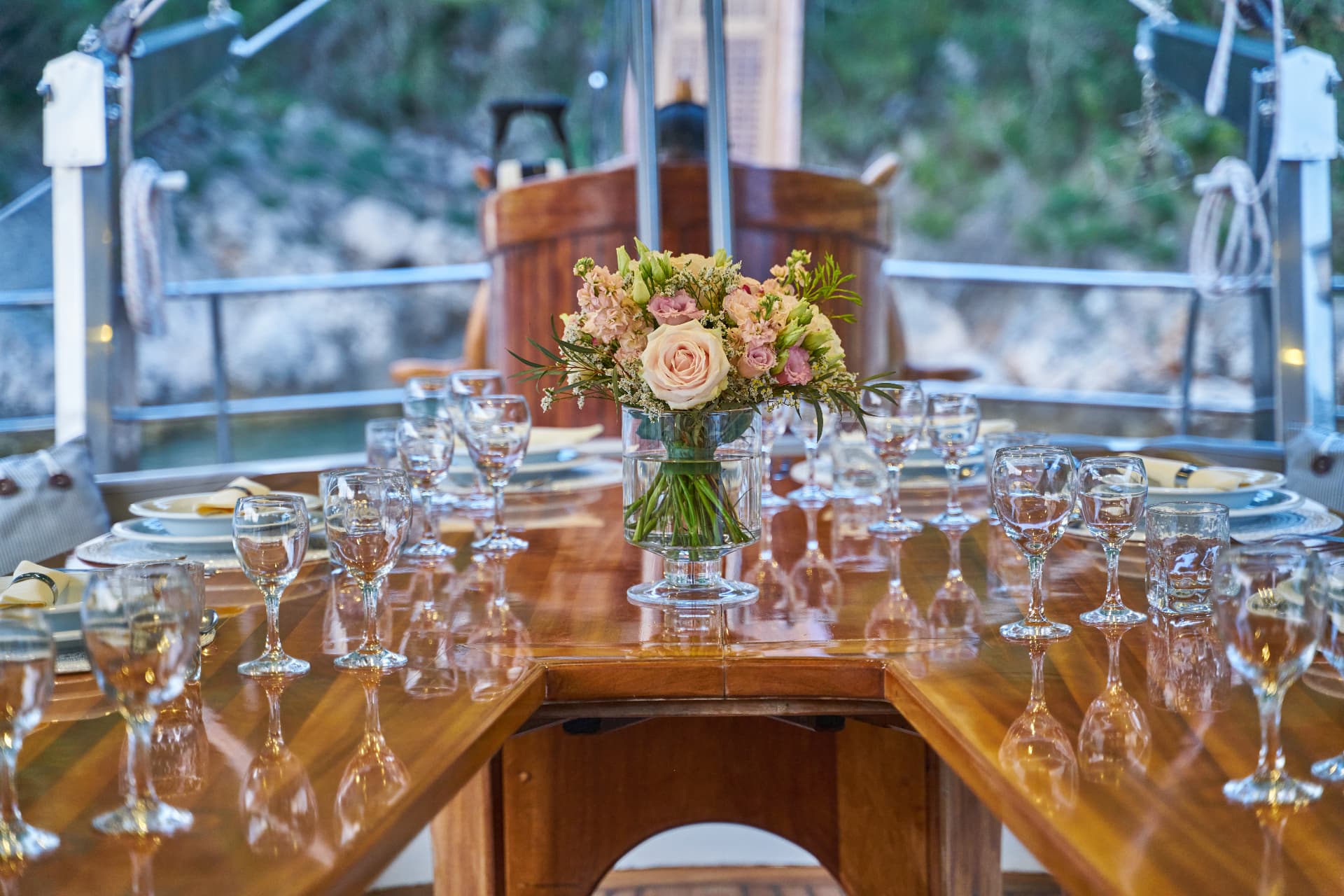 Formal dining table set on a boat deck with a floral centerpiece and blurred greenery outside.