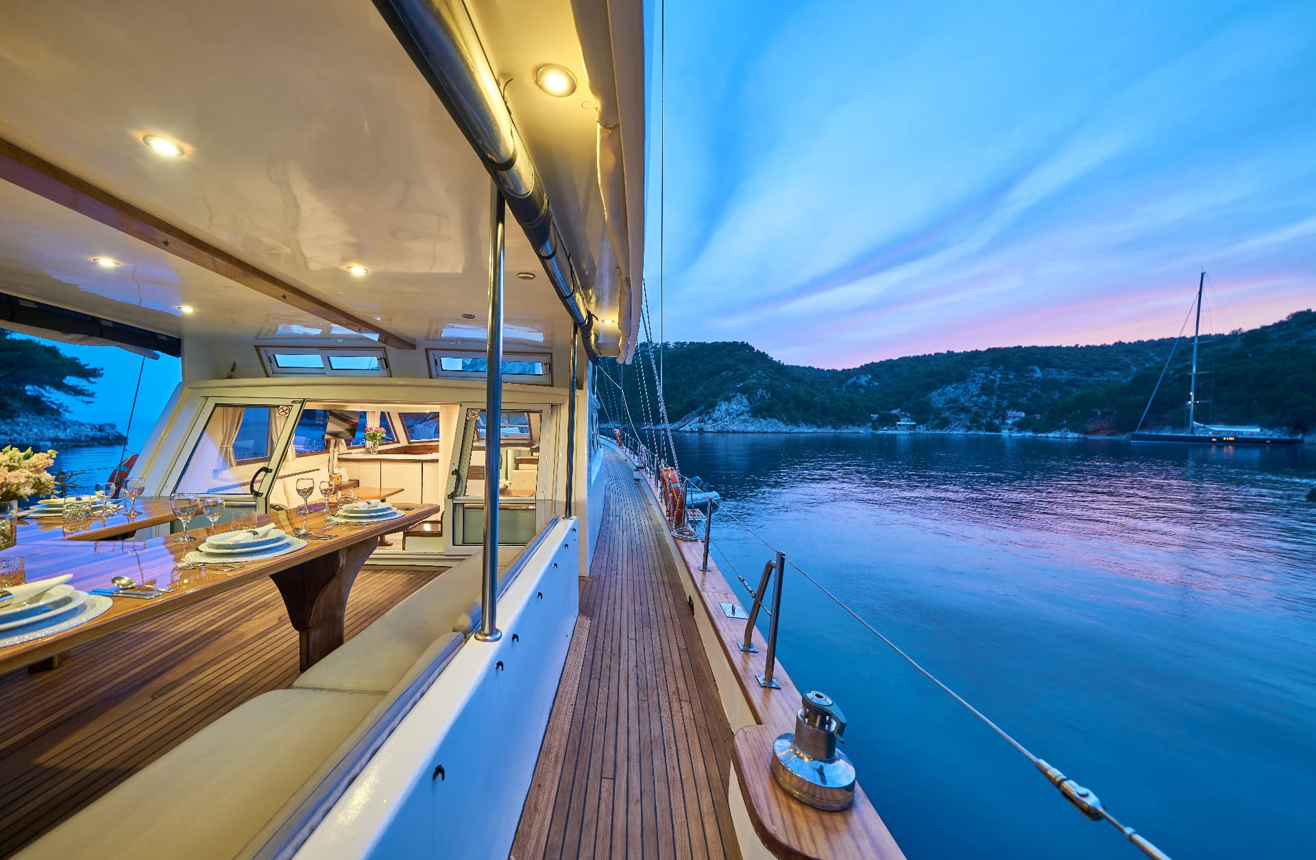 Yacht deck dining area illuminated at dusk near wooded coastline with anchored sailboat.