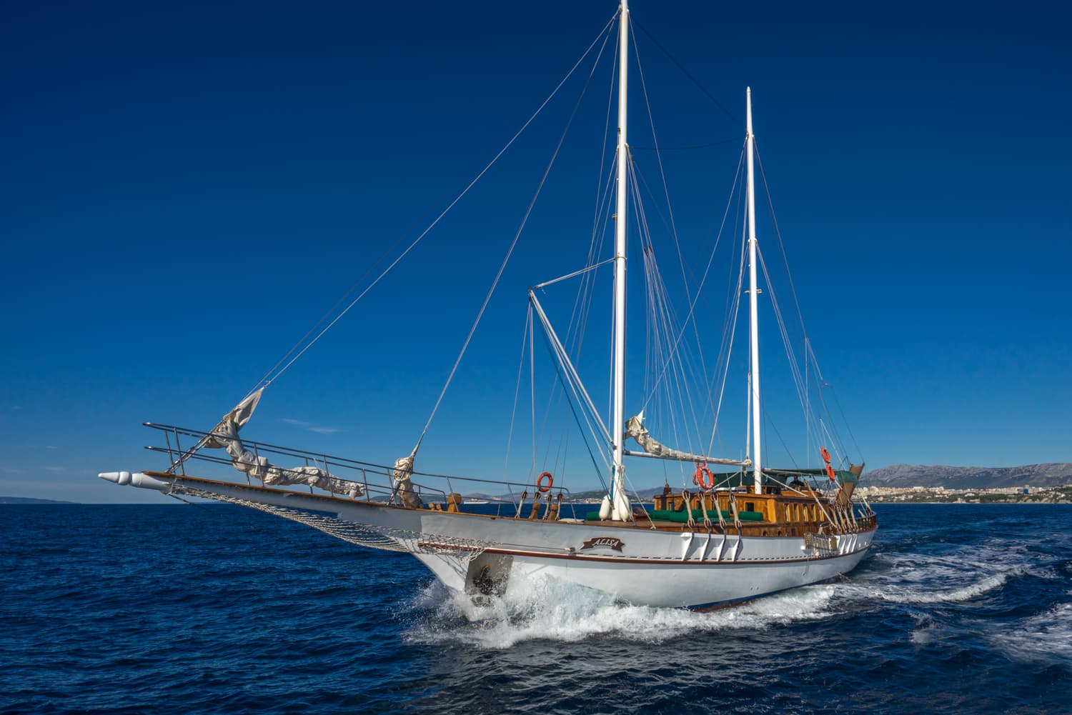 White sailboat named Alisa moving through deep blue water with distant coastline and mountains.