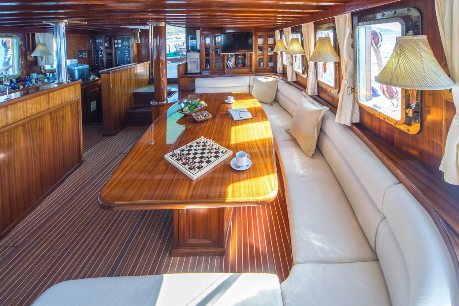 Yacht interior saloon with glossy wood, white seating, and a chessboard on the dining table.