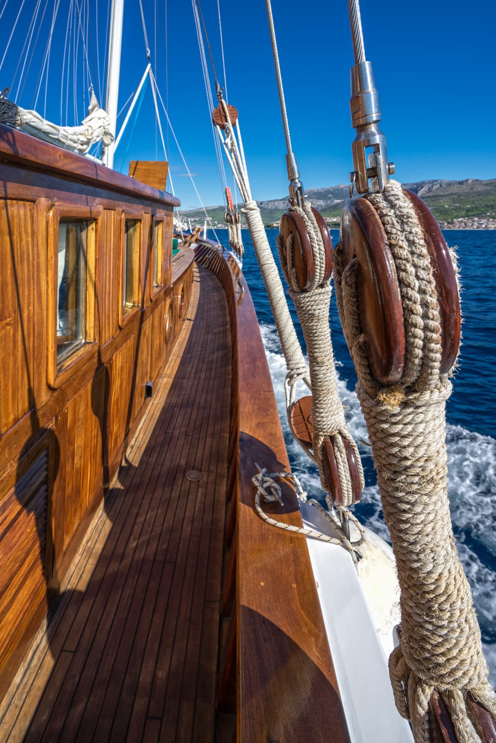 Wooden sailboat deck and rigging moving through blue sea toward distant coastline under clear sky.