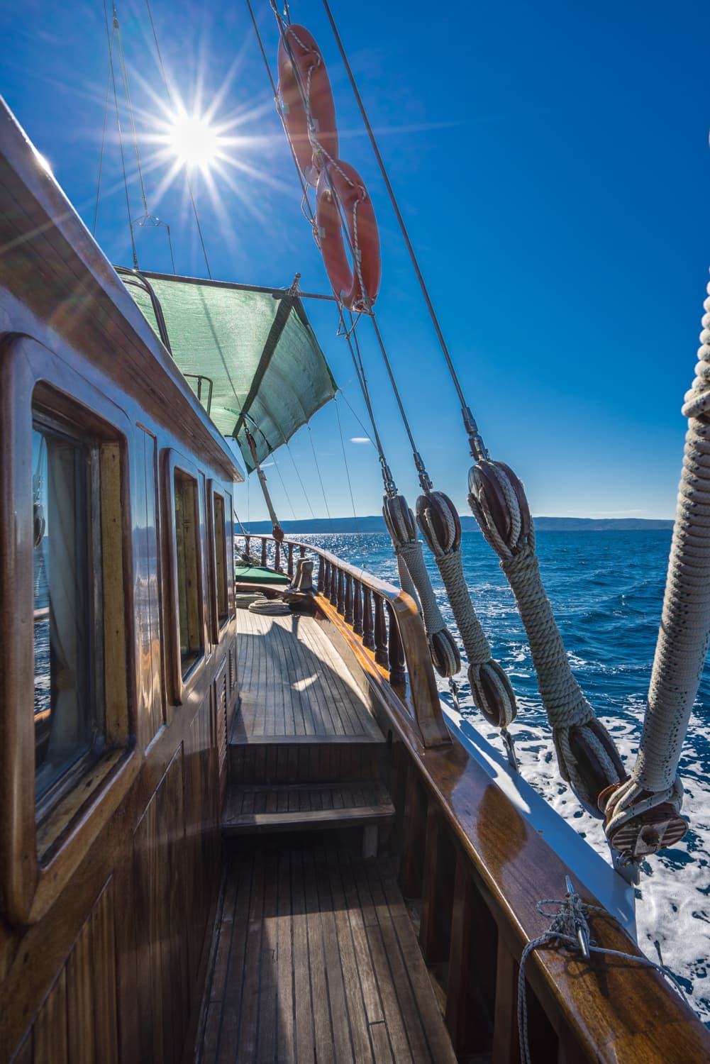 Wooden sailboat deck with rigging, life preservers, and bright sun over blue sea.