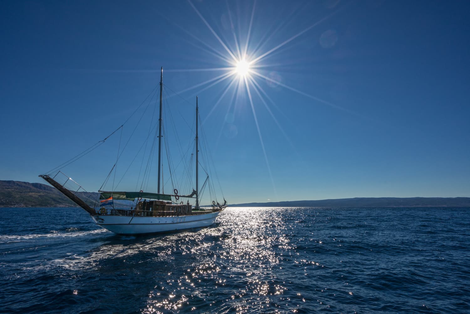 White sailboat cruising on deep blue sea under bright sun with distant coastline.