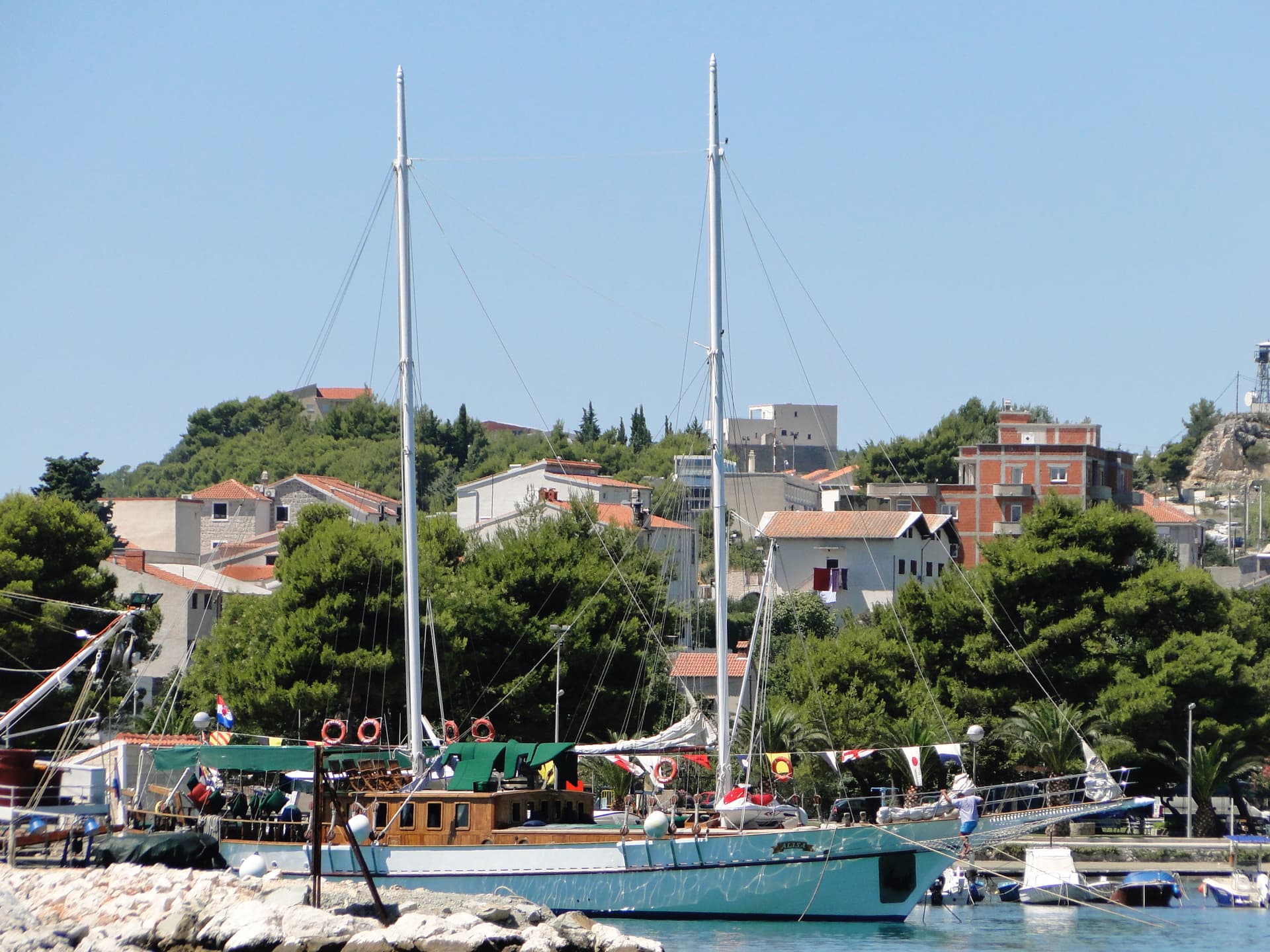 Two-masted sailboat named Alisa docked near a rocky shore with hillside homes and green trees.