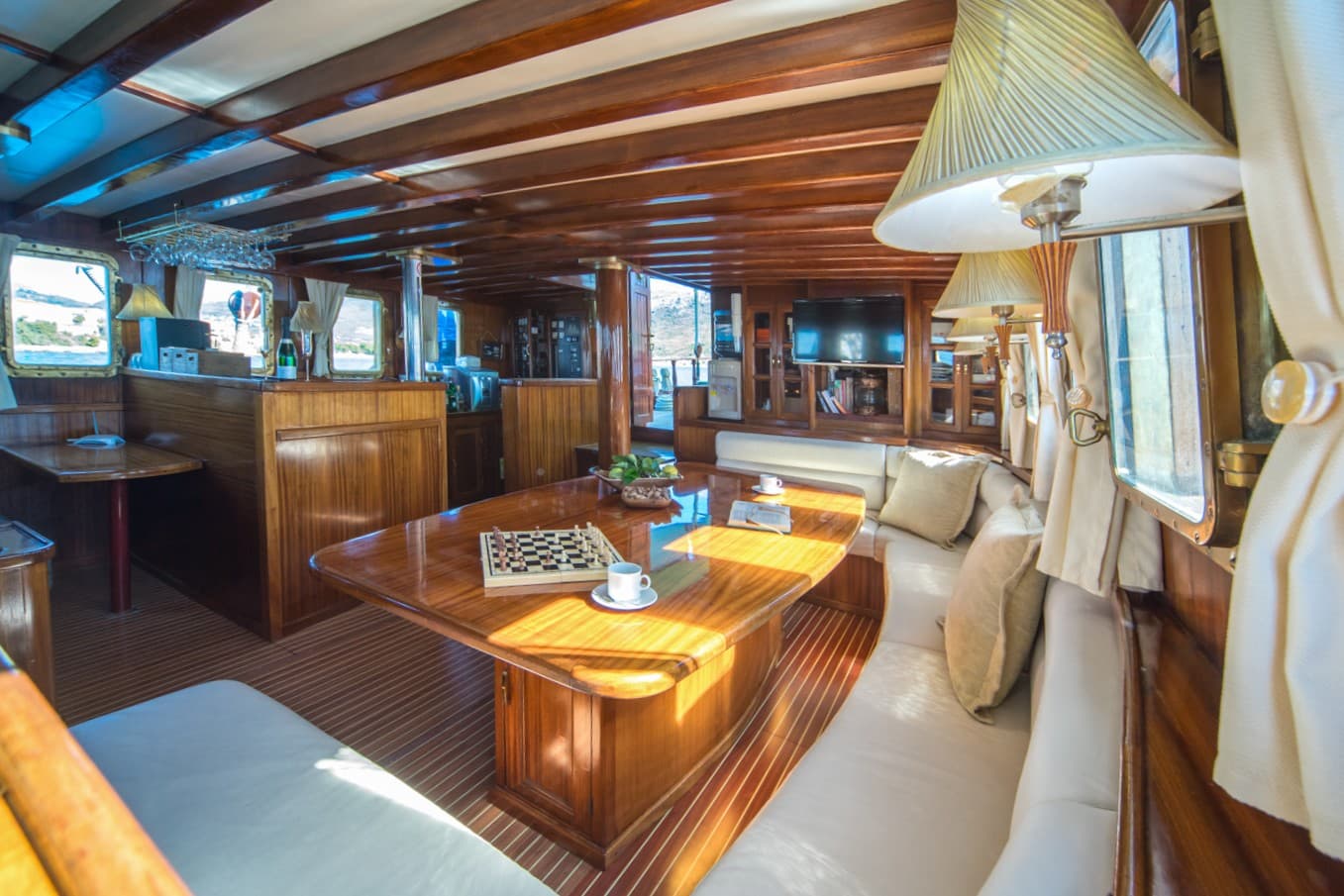 Interior cabin of a wooden yacht with a glossy table, chessboard, and white seating, showing a view outside.