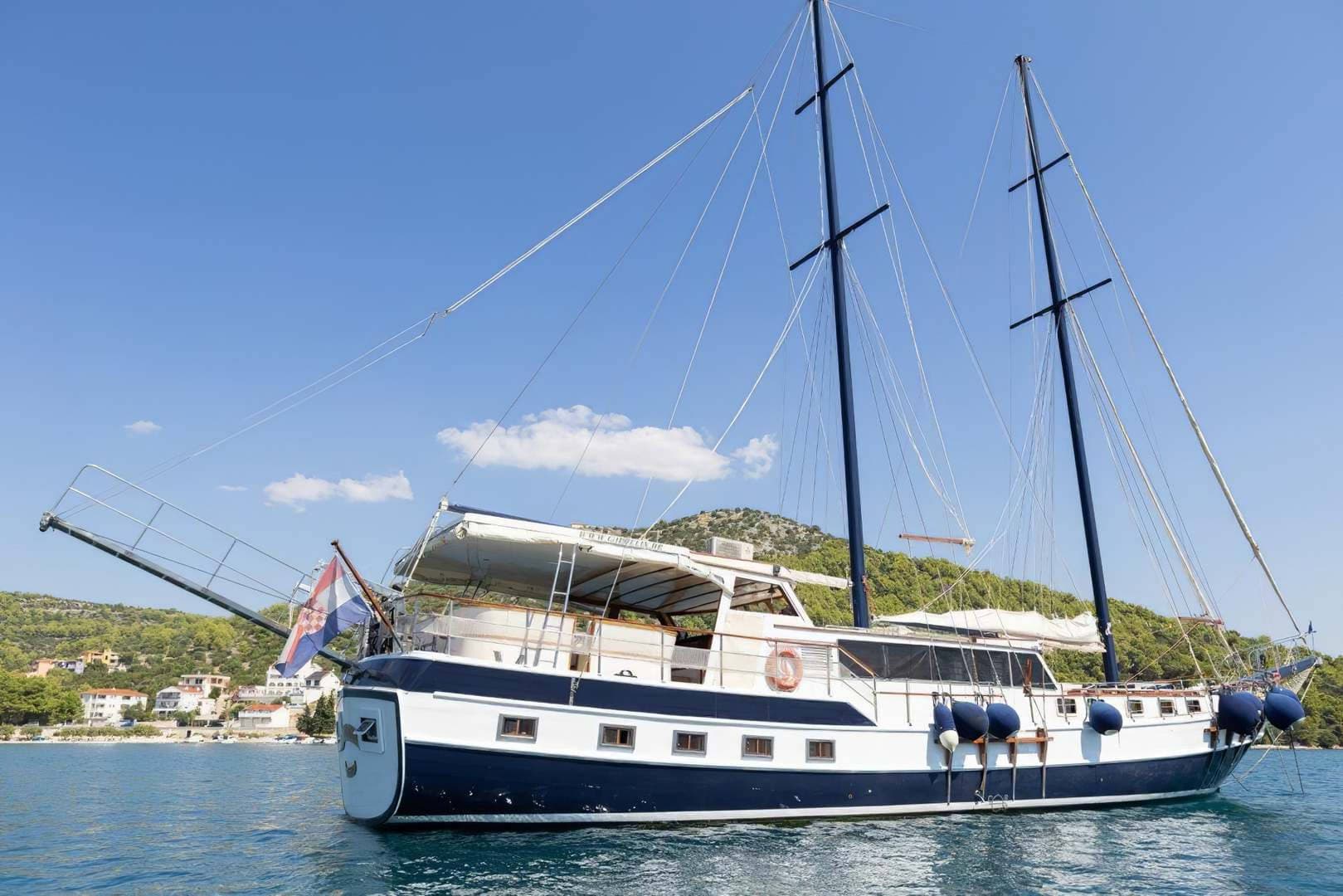 Blue and white sailboat with Croatian flag anchored near a green, hilly coastline.
