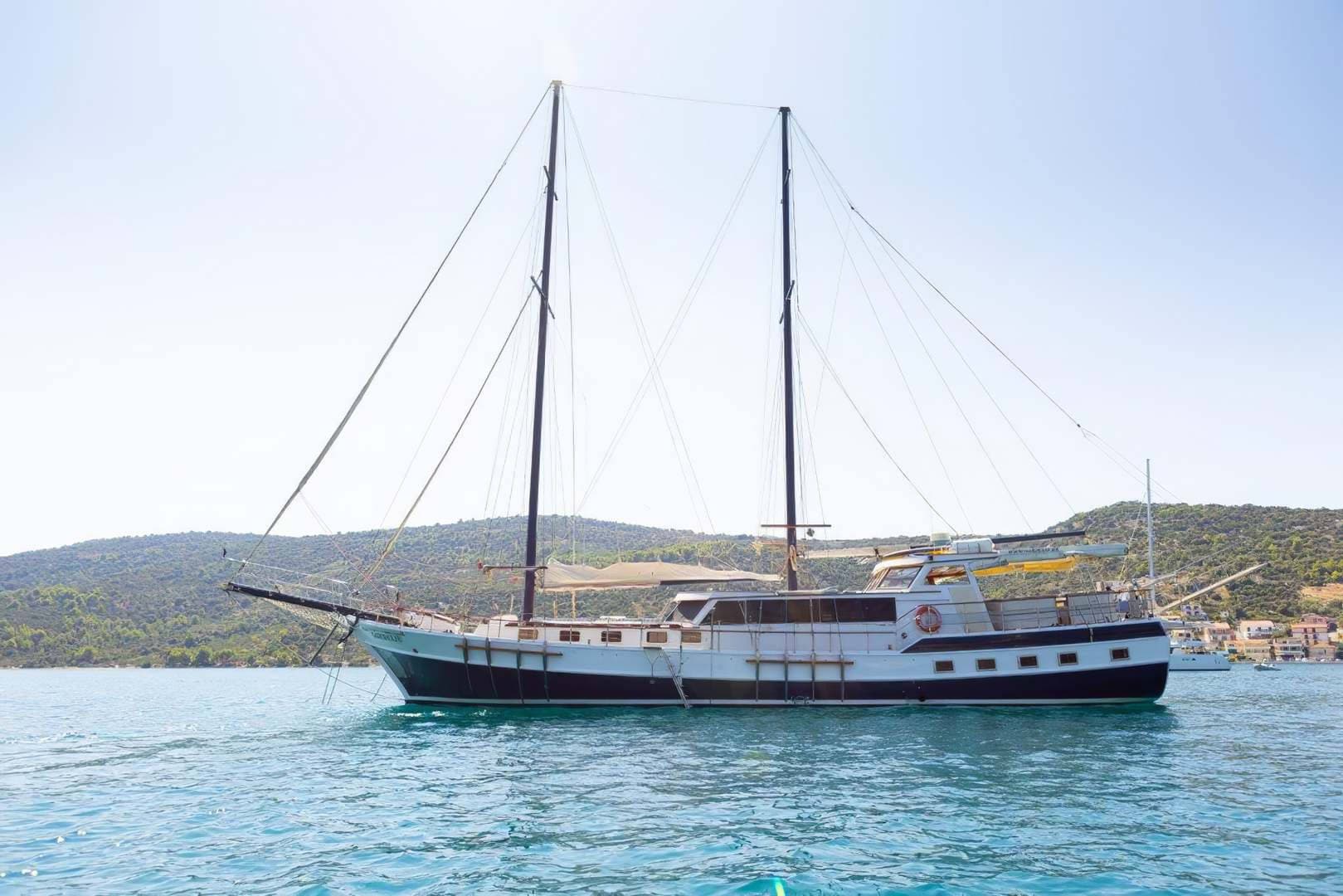 Two-masted wooden boat anchored in turquoise water near a tree-covered coastline.