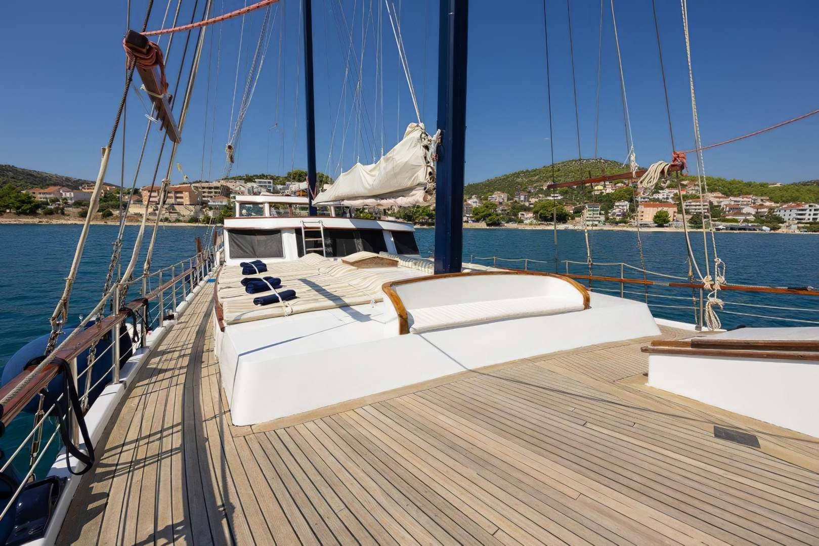 Wooden deck of a sailboat with rigging, overlooking a coastal town on a sunny day.