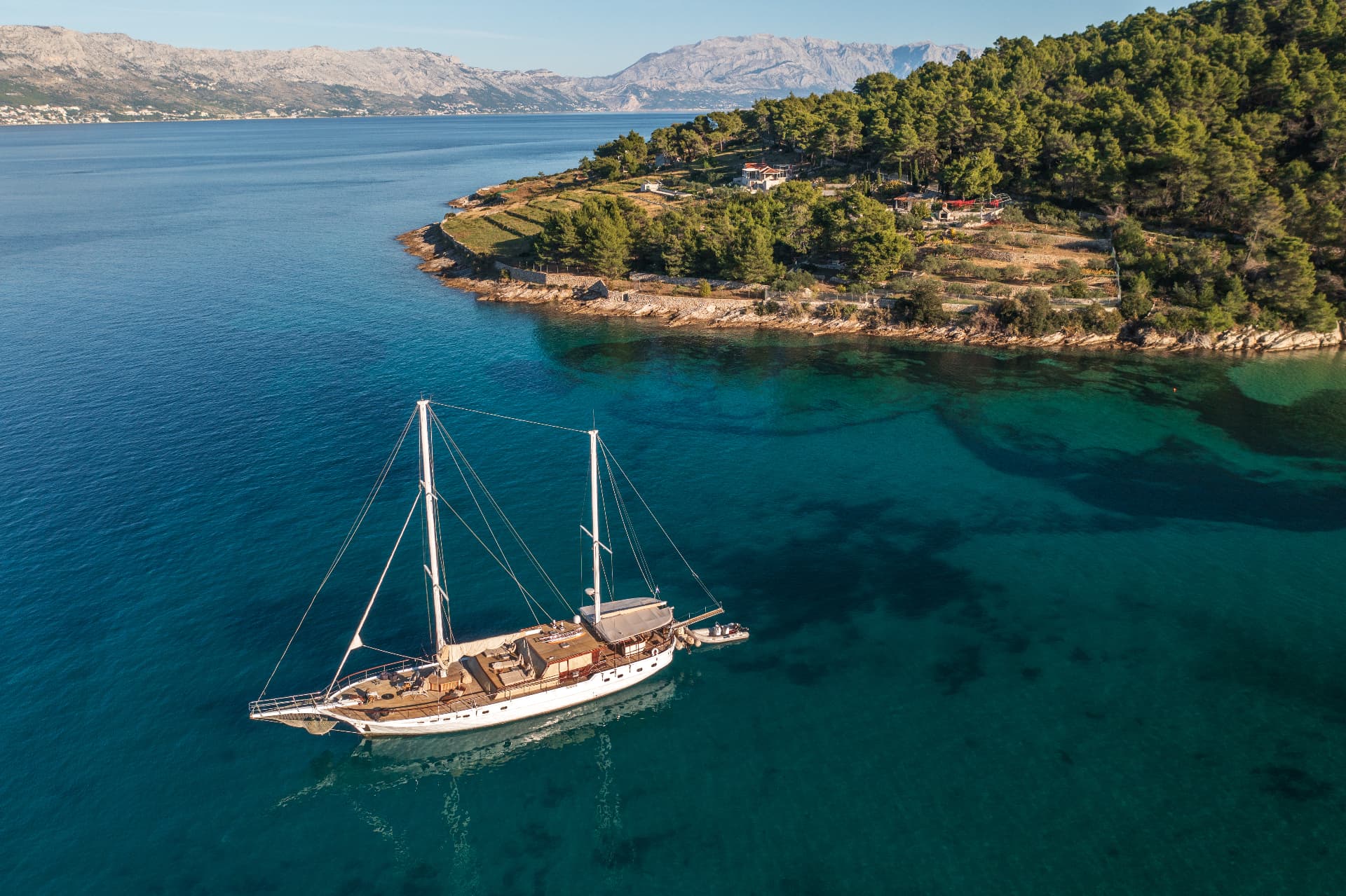 Sailboat anchored in clear turquoise cove near forested coastline and distant mountains.