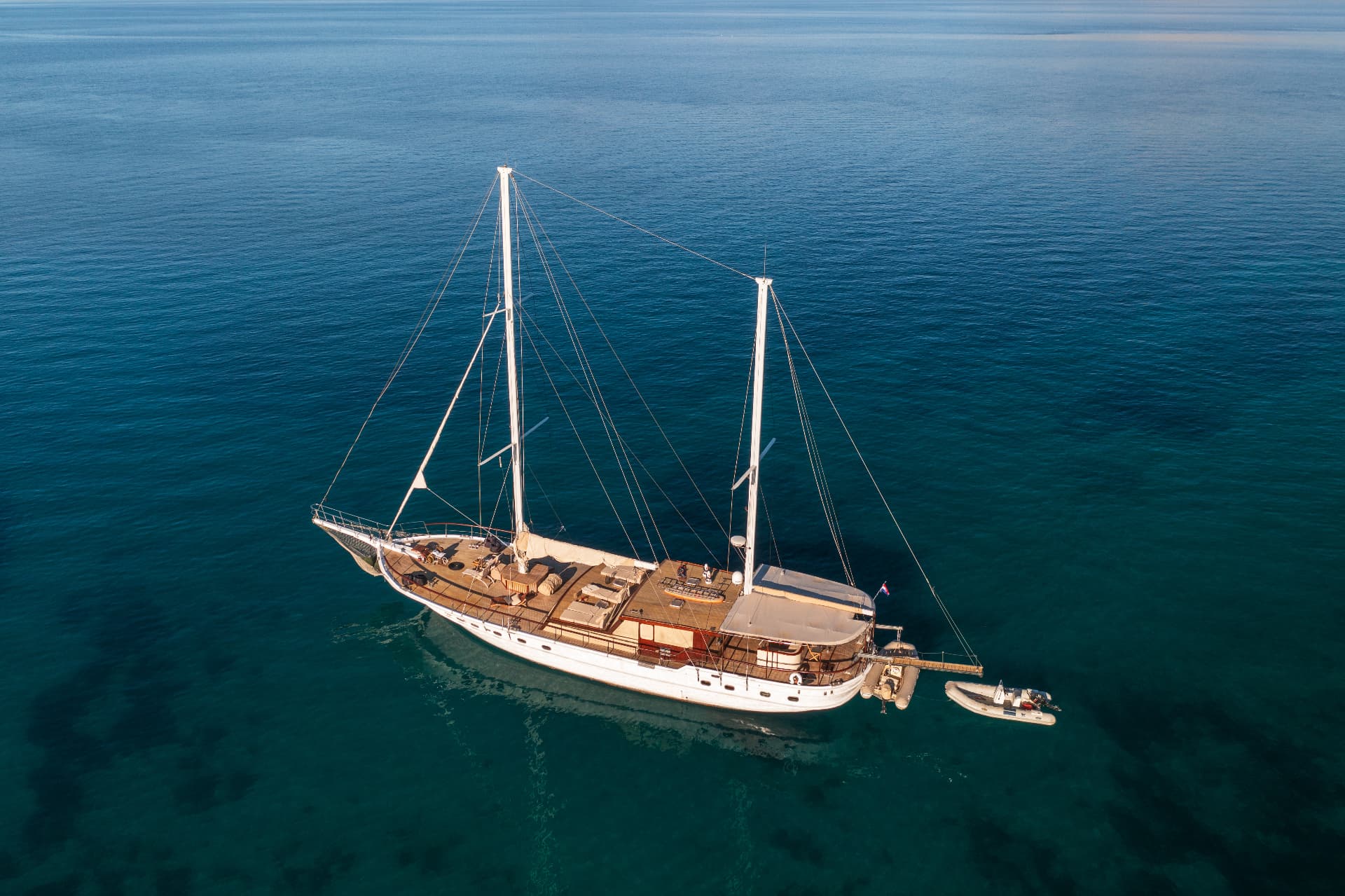 Large white sailboat anchored on deep blue, clear coastal water with a small dinghy attached.