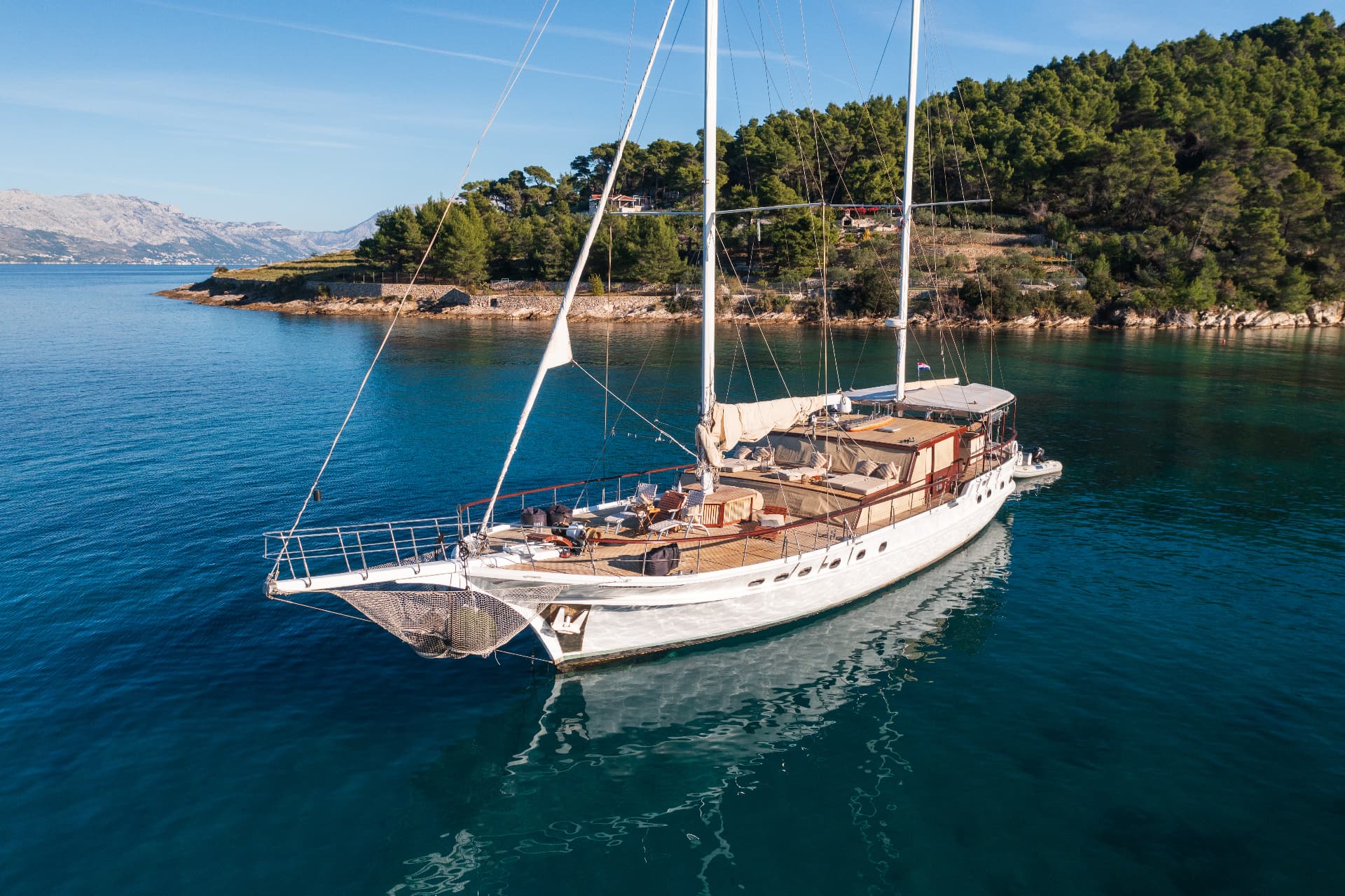 Large white sailboat anchored in clear blue water near a forested coastline with distant mountains.