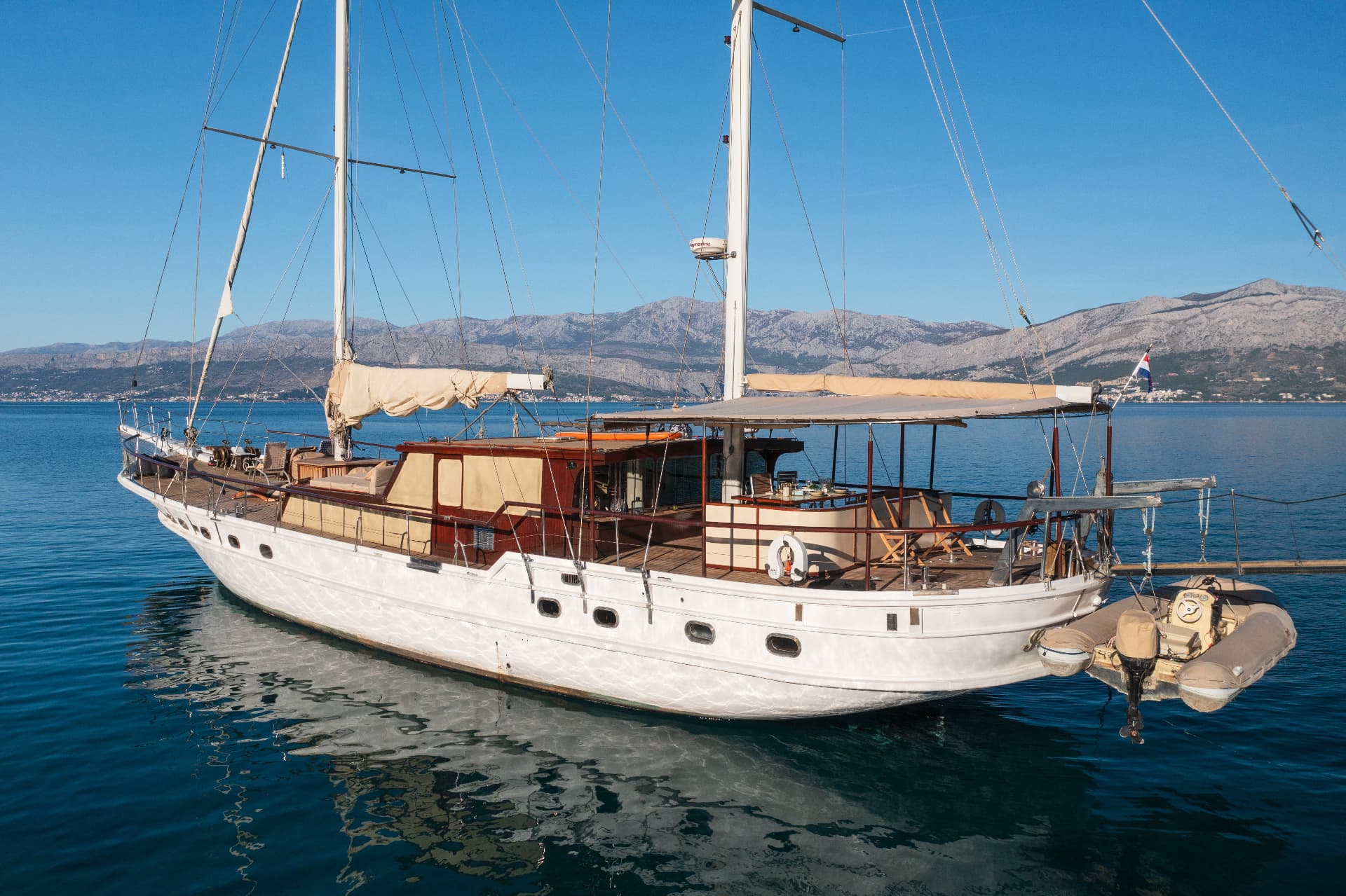 Large white sailboat with wooden cabin floating on calm blue water near mountains.