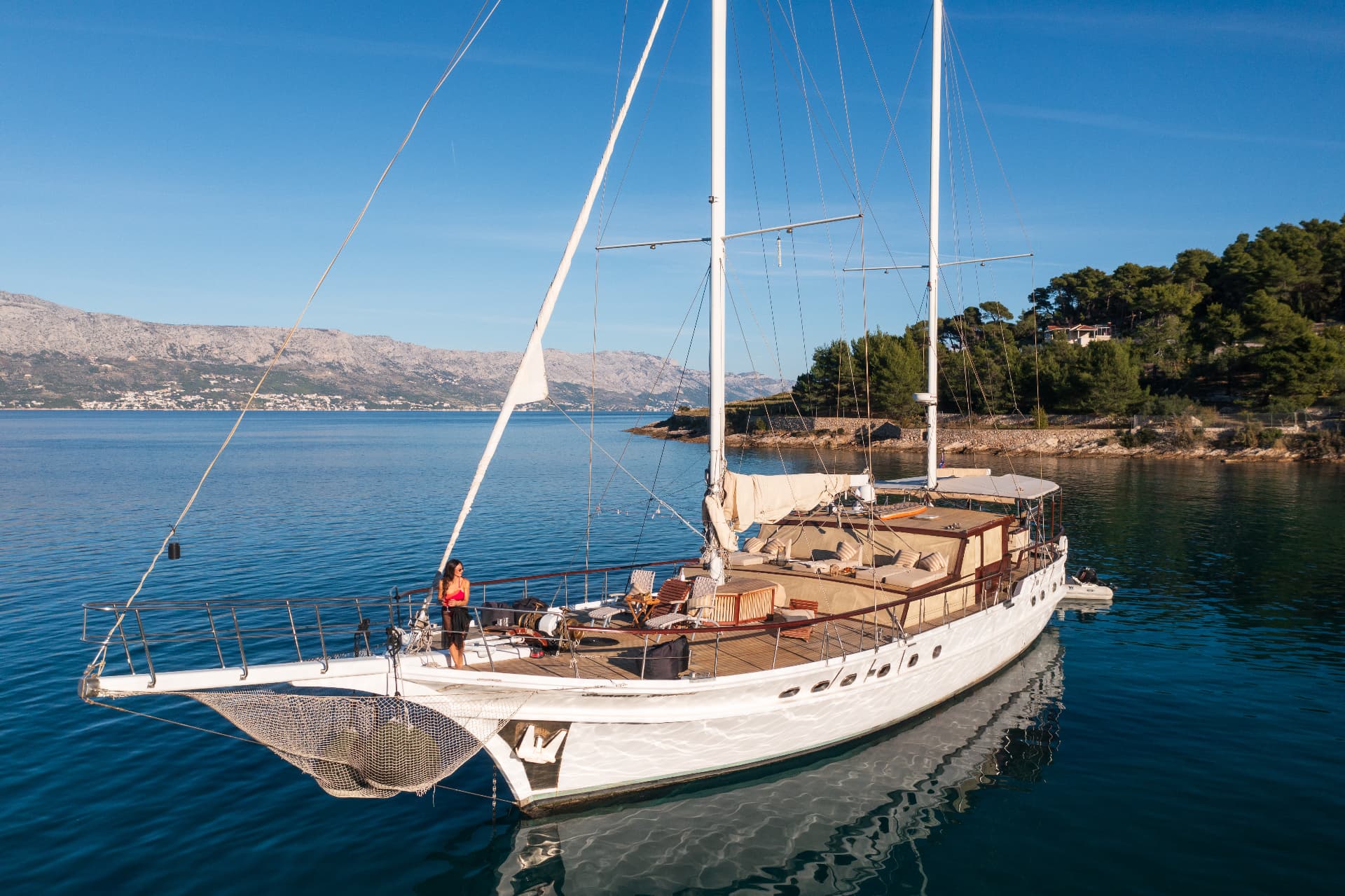 Large sailboat anchored in calm blue water near a wooded coastline with mountains across the bay.