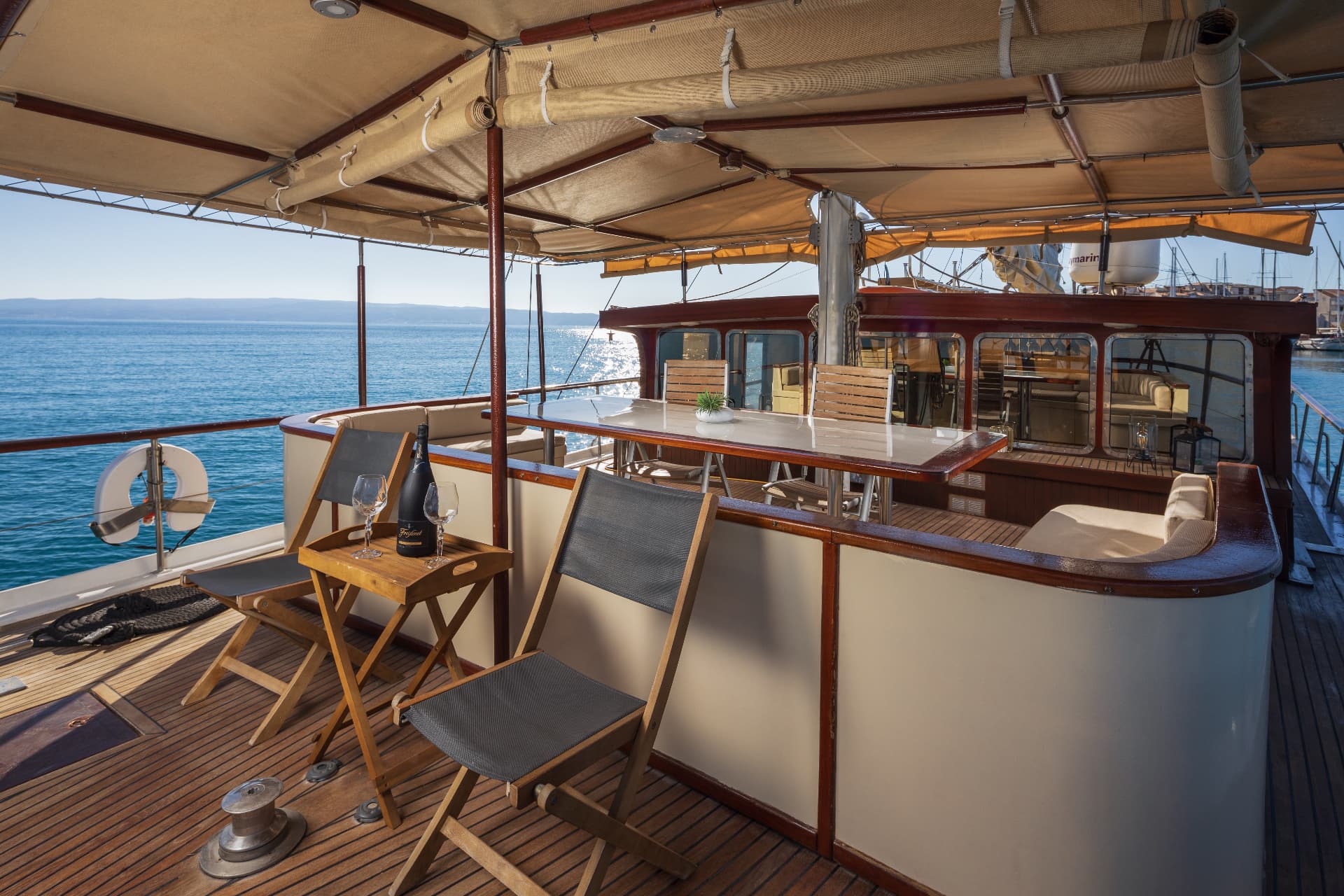 Boat deck with seating, champagne, and sea view under canvas awning in harbor.