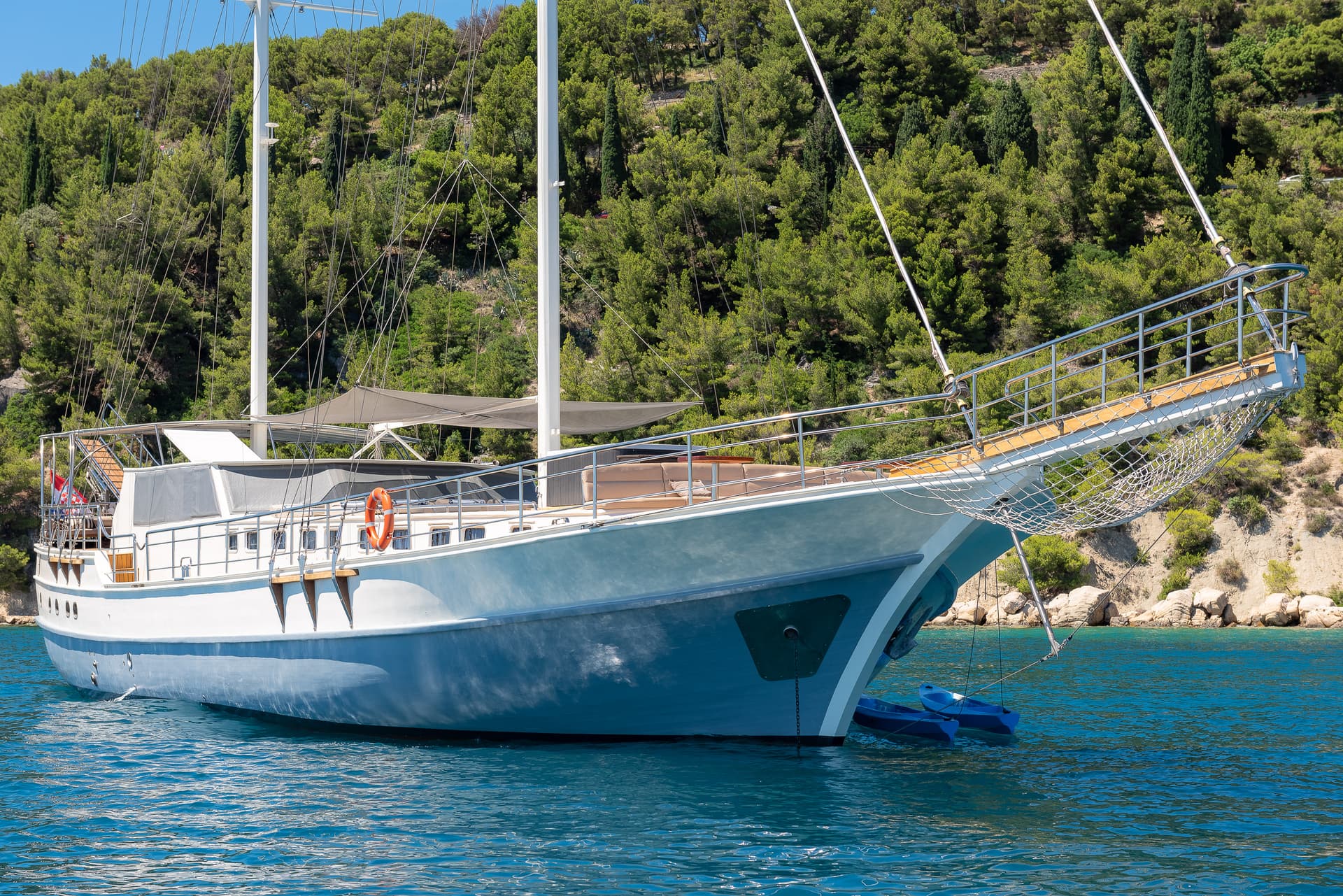 Large sailboat with white masts floating on blue water near a densely forested coastline.