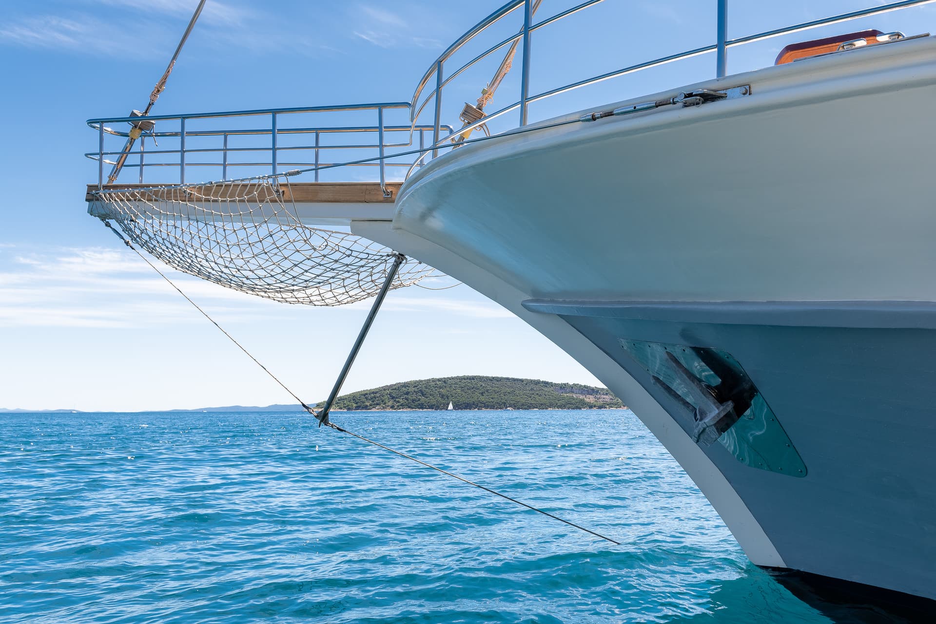 Bow of a boat with a bowsprit net over bright blue water near a forested island.