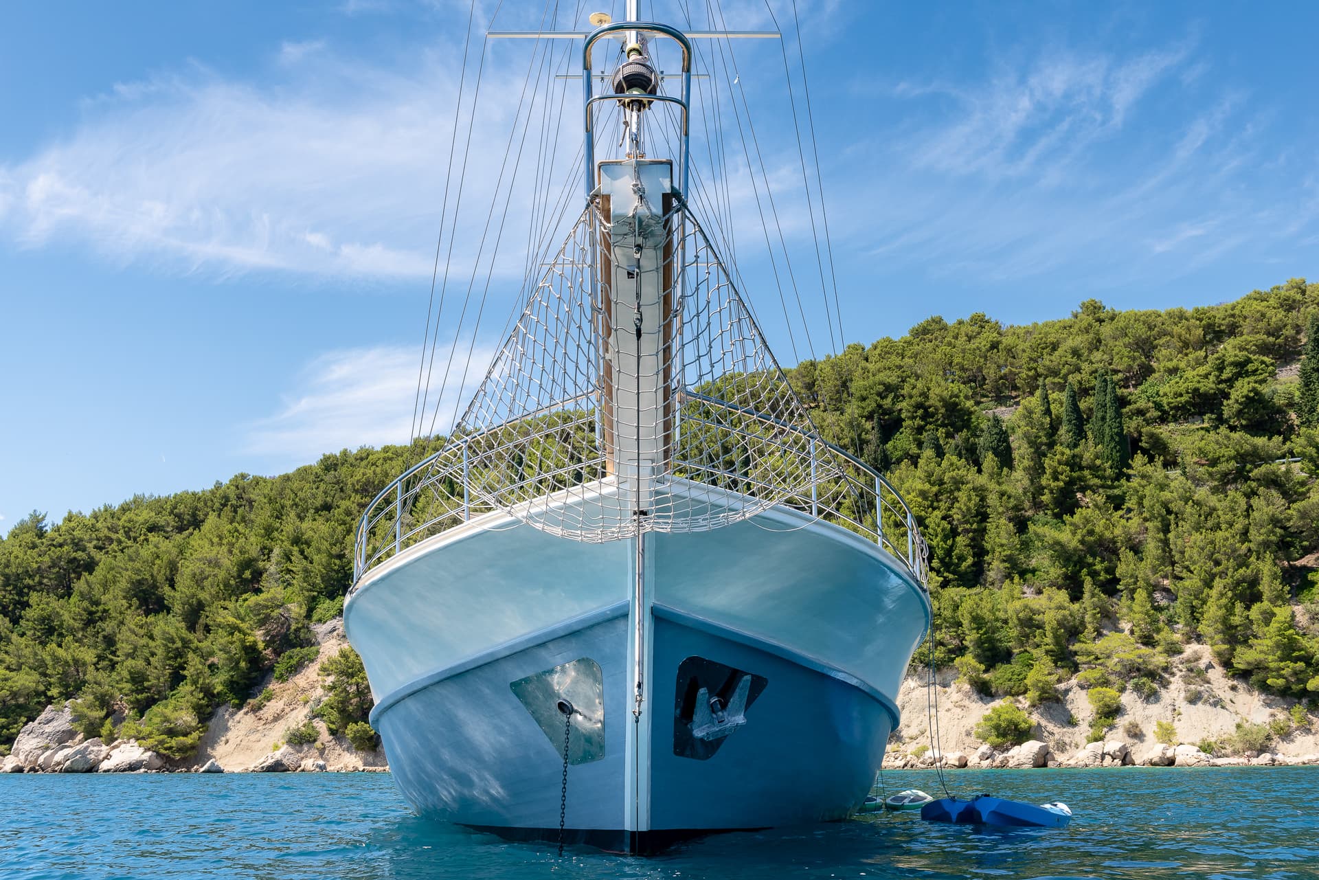 Boat bow with netting and anchor chain on blue water near forested rocky coastline.