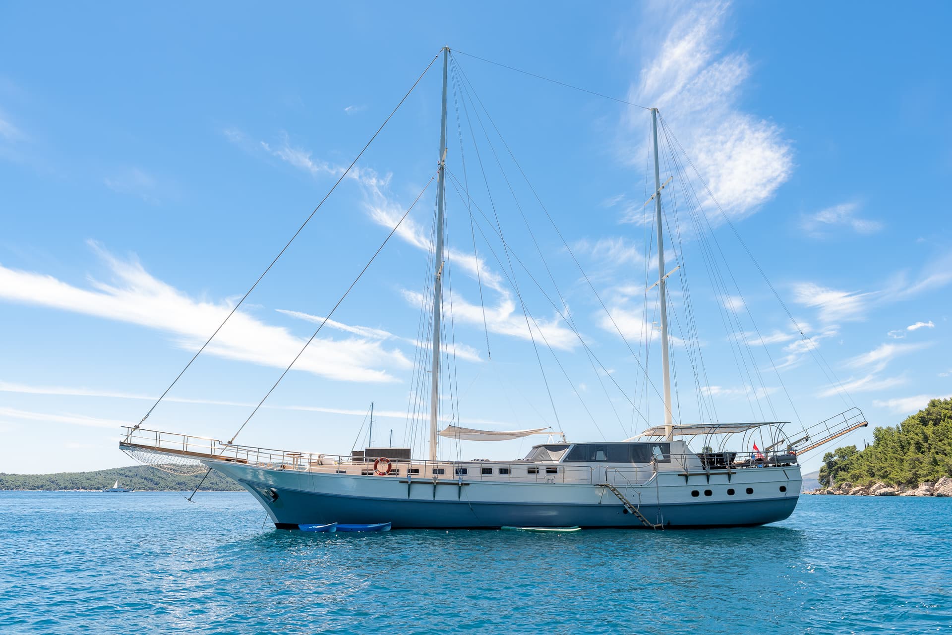 Large sailboat anchored on blue water near a wooded coastline under a bright blue sky.