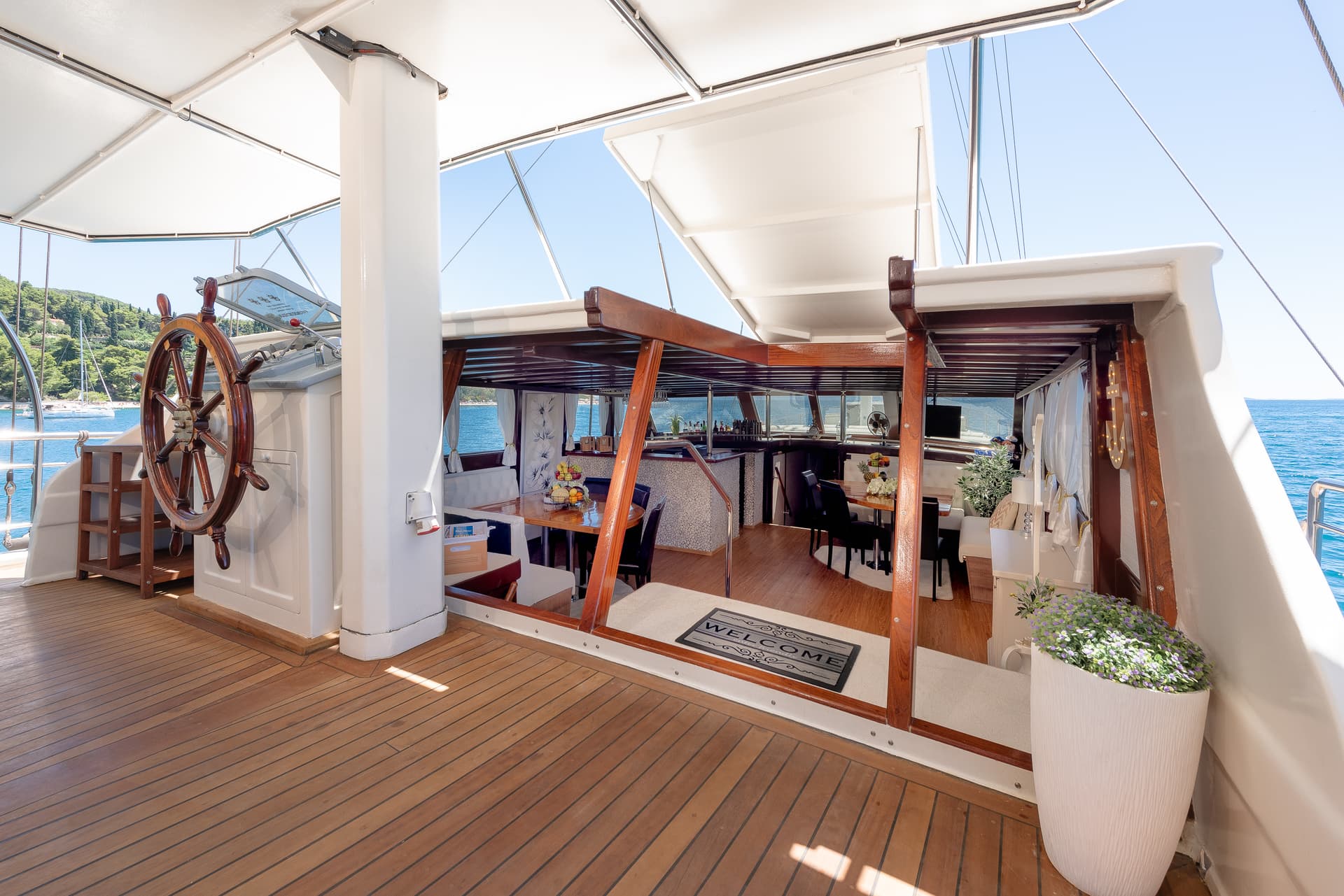 Boat deck with wooden helm, dining area, and lush green coastline visible.