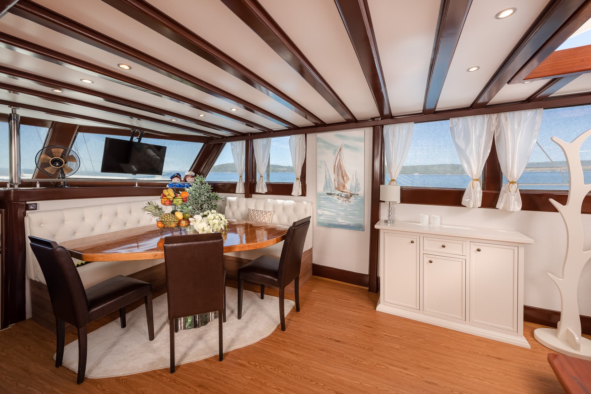 Yacht dining area with wooden table, white seating, and view of blue water and hills.
