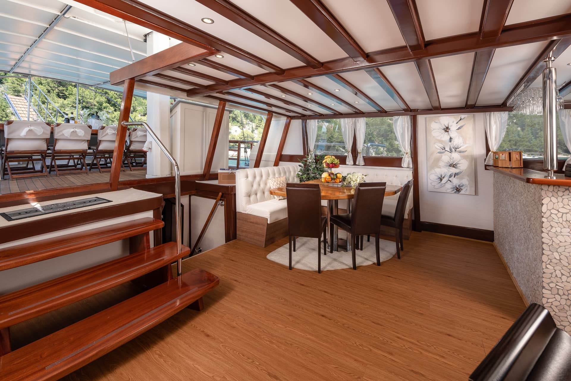 Interior dining area of a boat with wood trim, white seating, and view of green coastline.