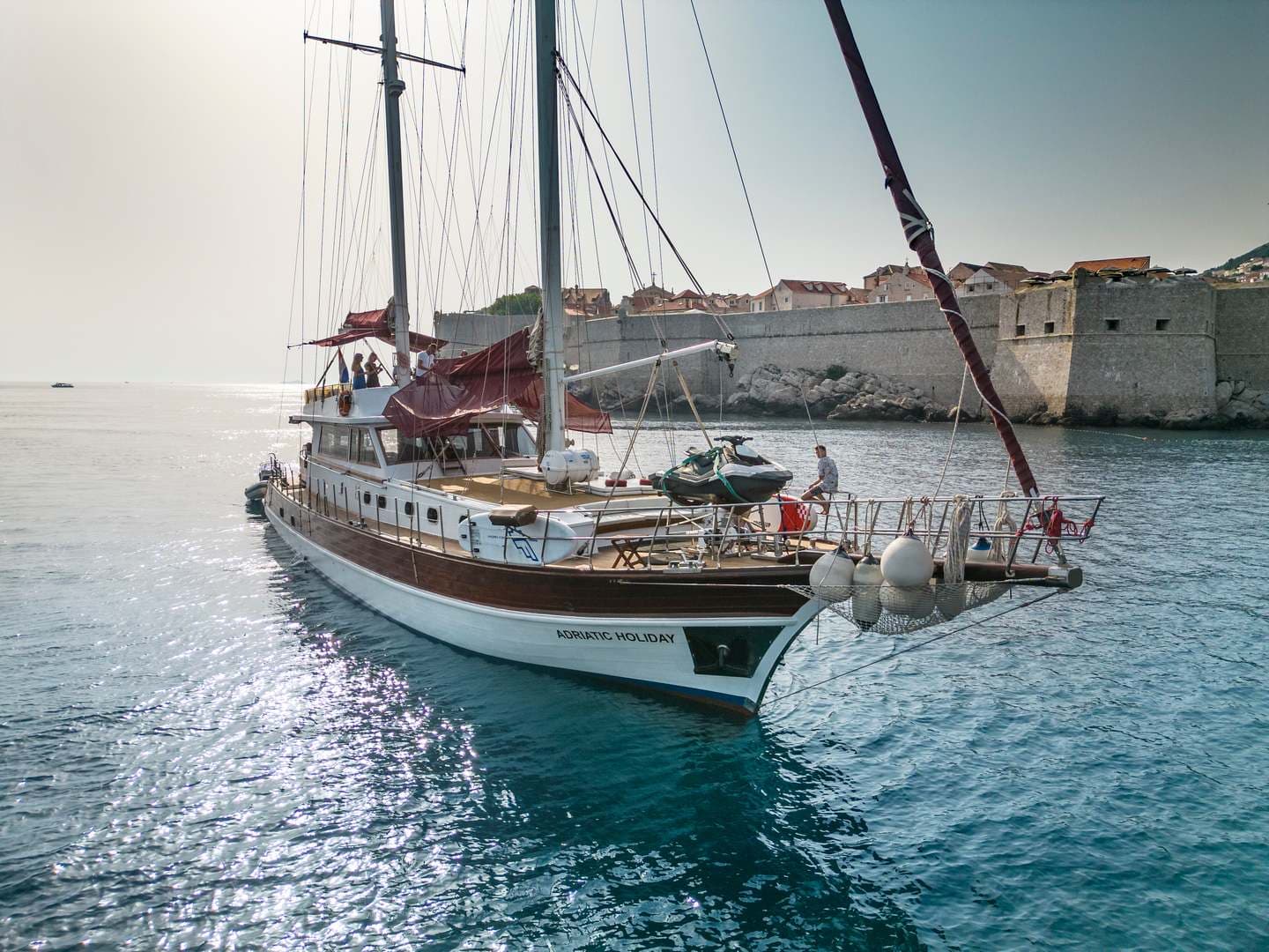 Large wooden sailboat named Adriatic Holiday near historic stone city walls on blue water.