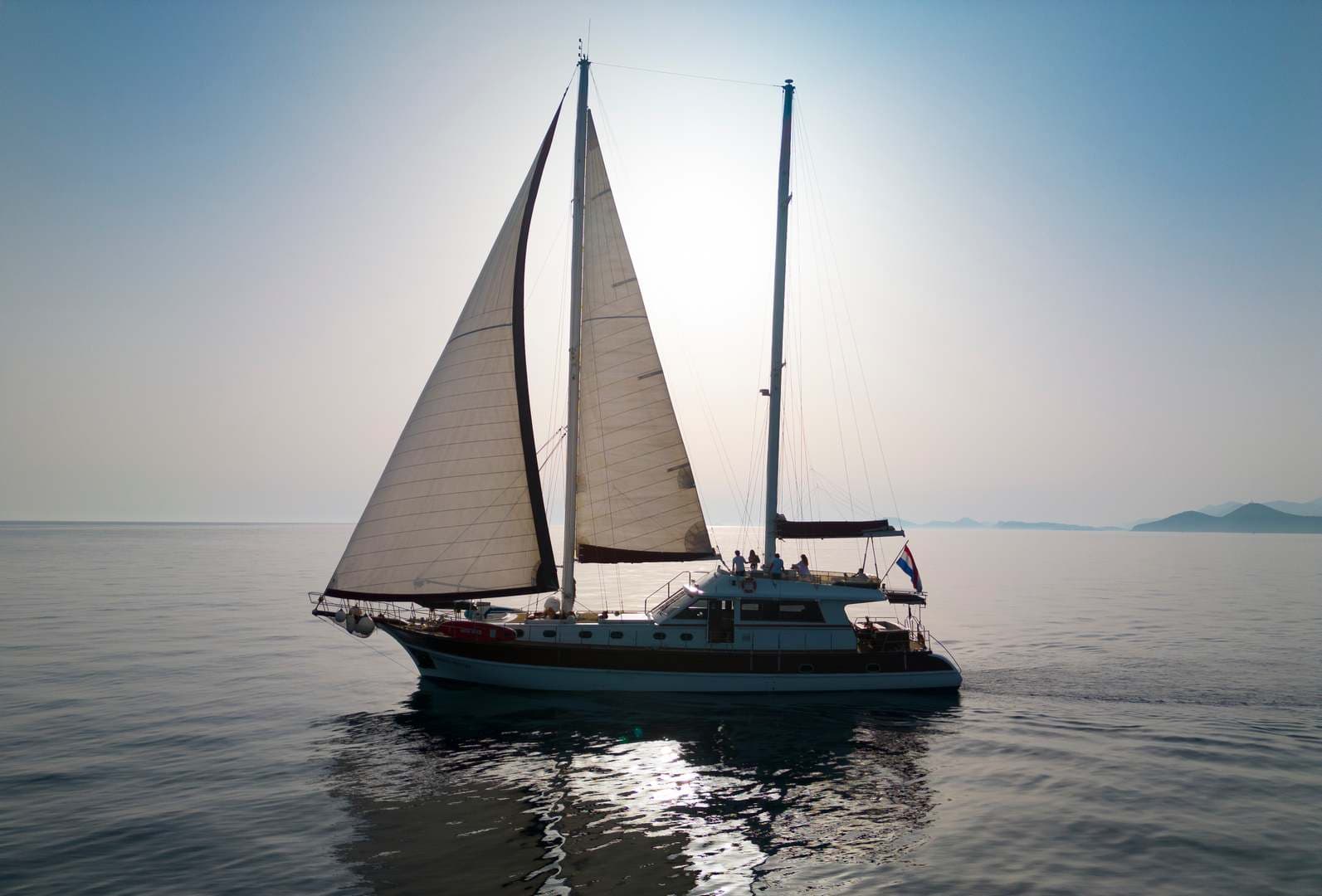 Sailboat with full sails on calm sea with distant mountains, Croatian flag visible.