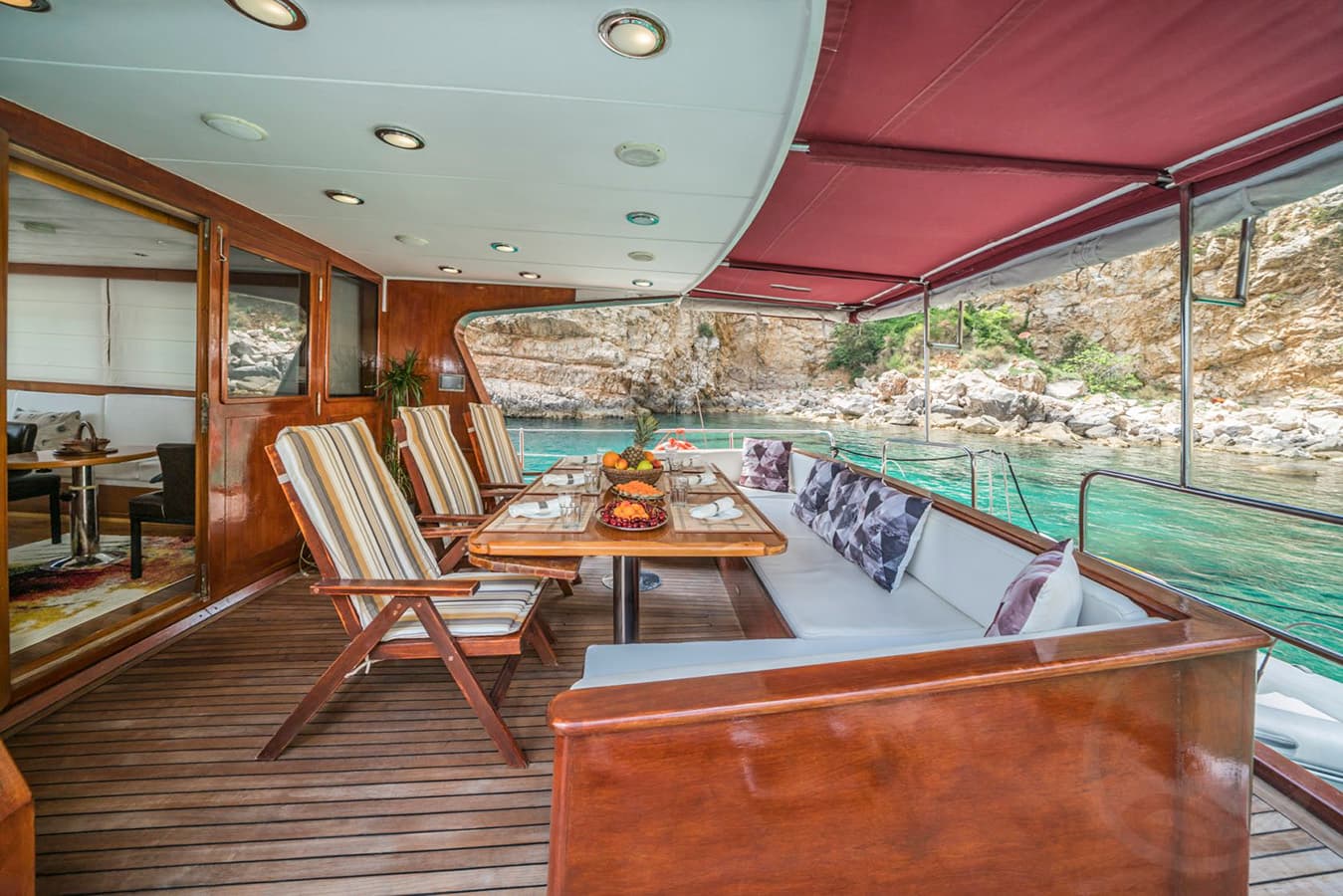 Yacht dining area with fruit bowl, anchored near rocky coastline with turquoise water