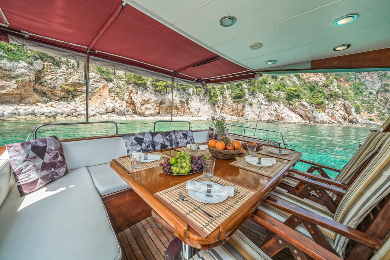 Boat dining area with fruit setting near rocky, green coastline and turquoise water