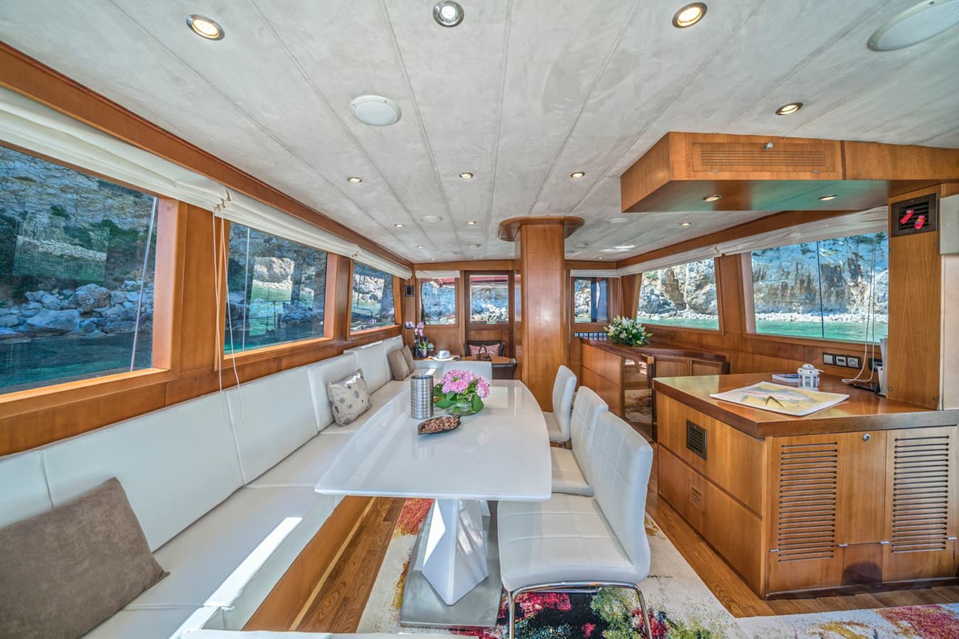 Yacht interior dining area with white seating, wood trim, and views of rocky coastline.
